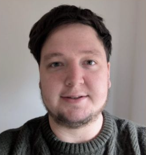 A young man with short dark hair, facial hair, and light skin taking a selfie indoors, wearing a gray sweater.