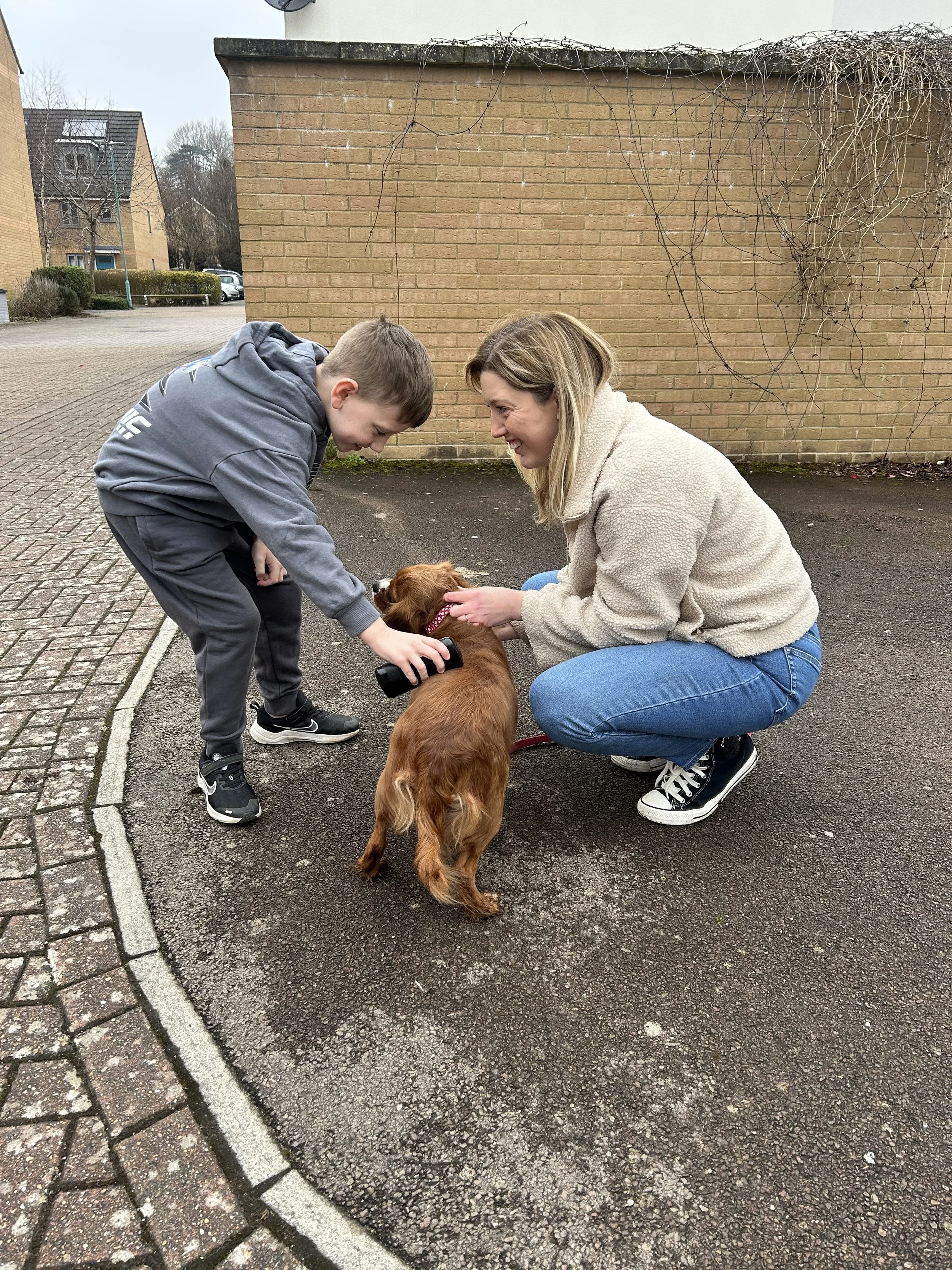 A woman and a boy are playing with a small brown dog outdoors on a paved surface. The woman crouches down, smiling and touching the dog's head, while the boy leans forward, holding a bottle near the dog's face. In the background, there is a brick wall and some leafless vines.