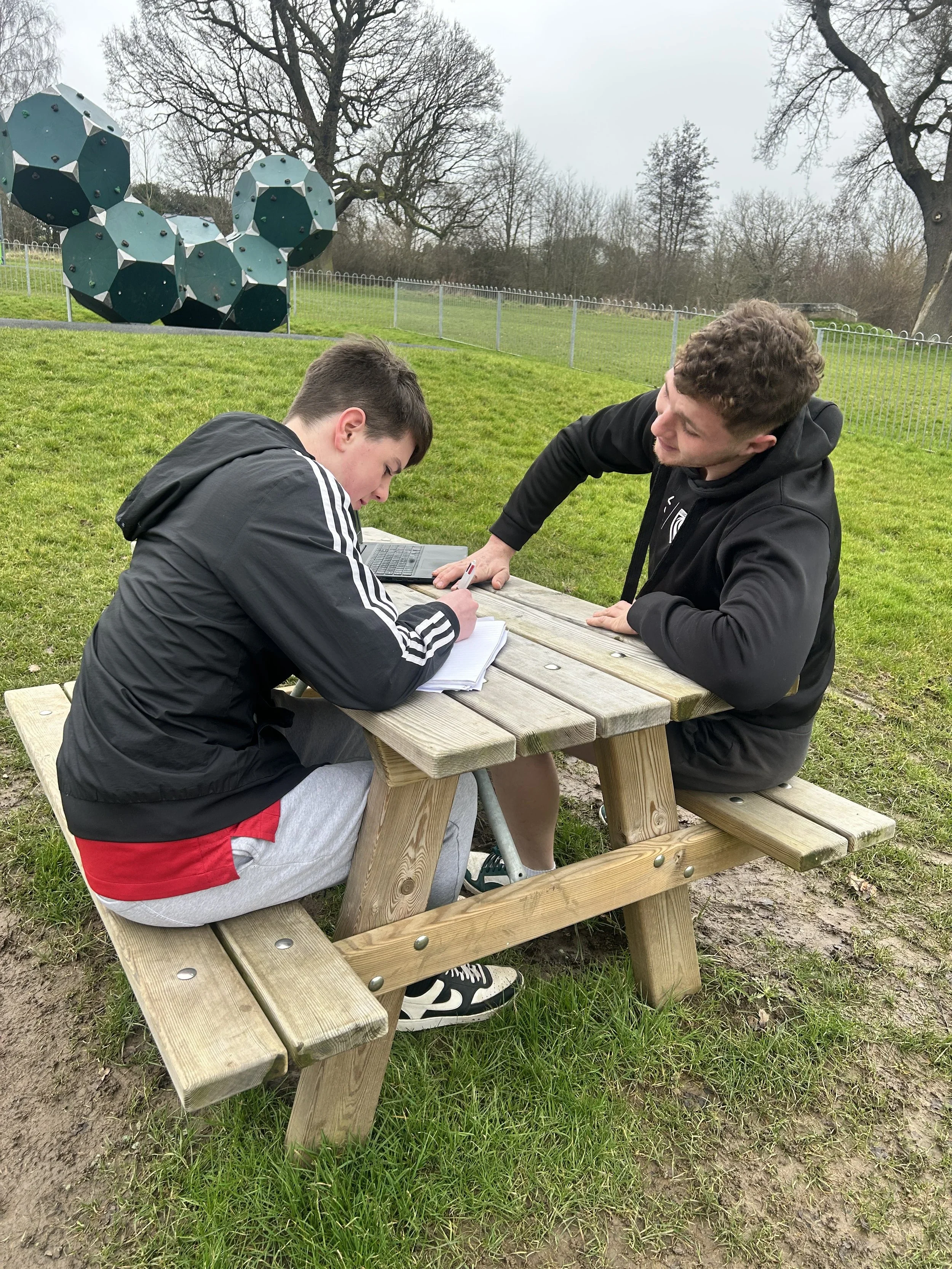 Two young men sitting at a wooden picnic table outdoors on a cloudy day, with grassy area and trees in the background, and a large geometric sculpture in the distance.