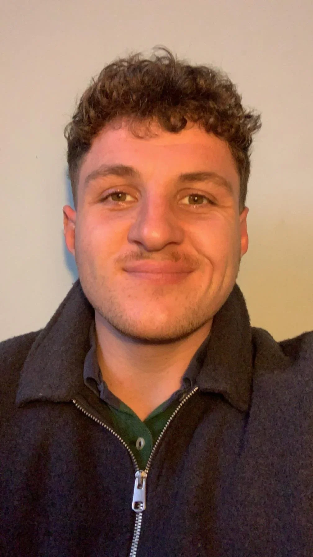 A young man with curly brown hair, light skin, and a slight beard, smiling, wearing a black zip-up jacket over a green collared shirt, standing against a plain wall.