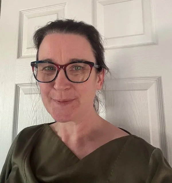A woman with dark hair, glasses, and a slight smile, standing in front of a white door.