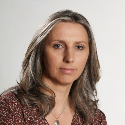 A woman with shoulder-length gray hair and blue eyes, wearing a patterned brown top with a necklace, looking directly at the camera against a plain light background.