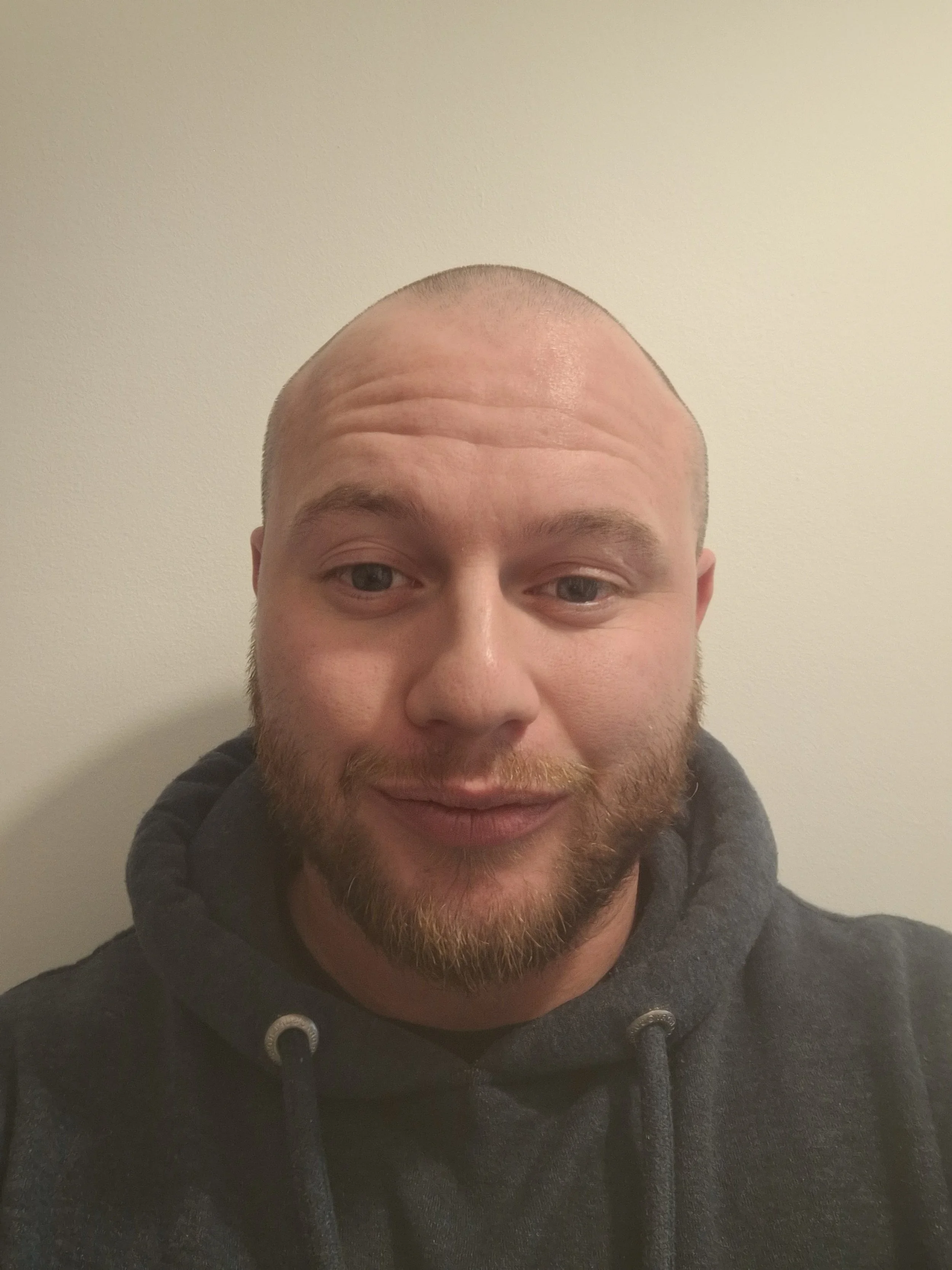 A man with a shaved head, a beard, and a mustache wearing a dark gray hoodie against a plain off-white wall