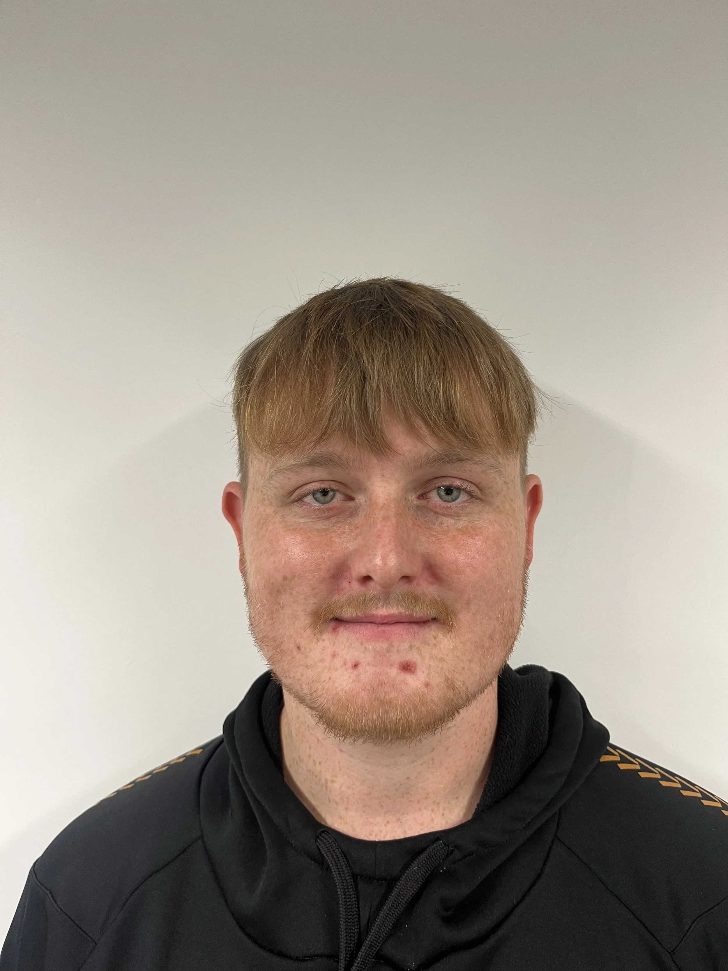 A young man with red hair and facial freckles wearing a black hoodie, standing against a plain light-colored wall.