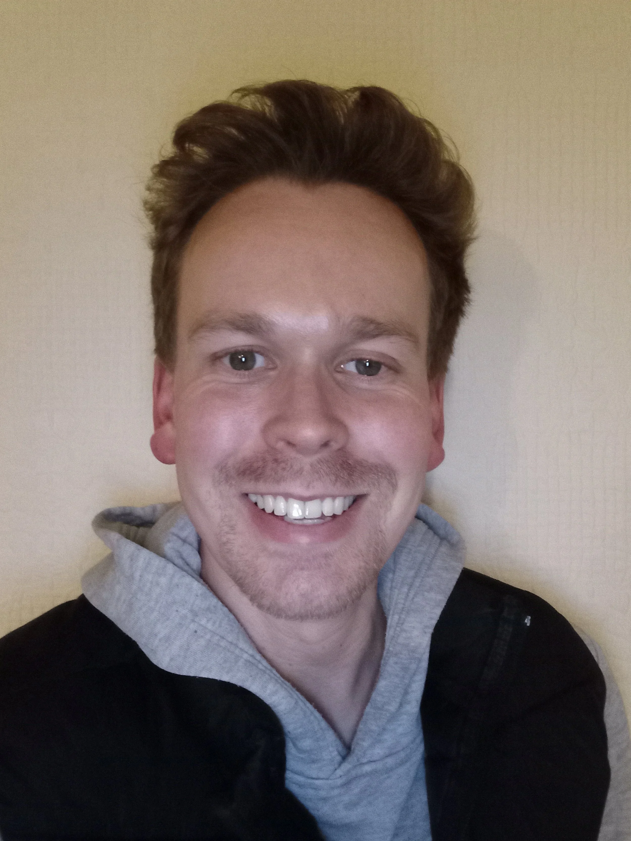 Smiling young man with short brown hair, wearing a gray hoodie and black jacket, standing against a beige wall.