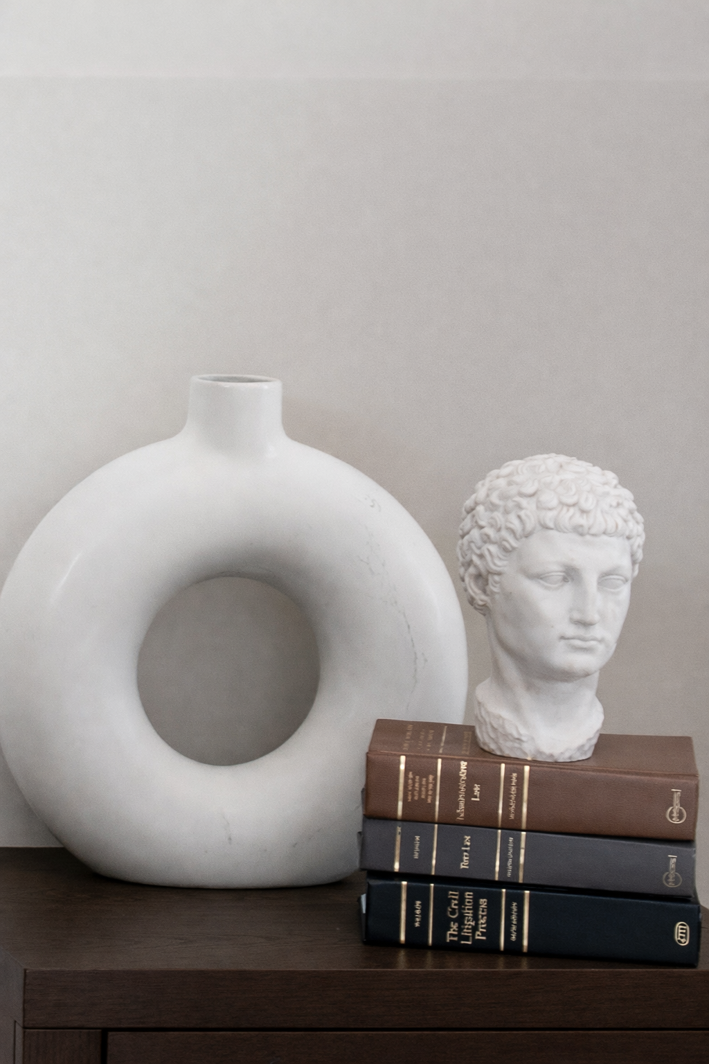 Decorative objects including a white ceramic vase with a hollow center, a white bust of a person's head, and a stack of four hardcover books on a dark wooden surface against a light-colored wall.