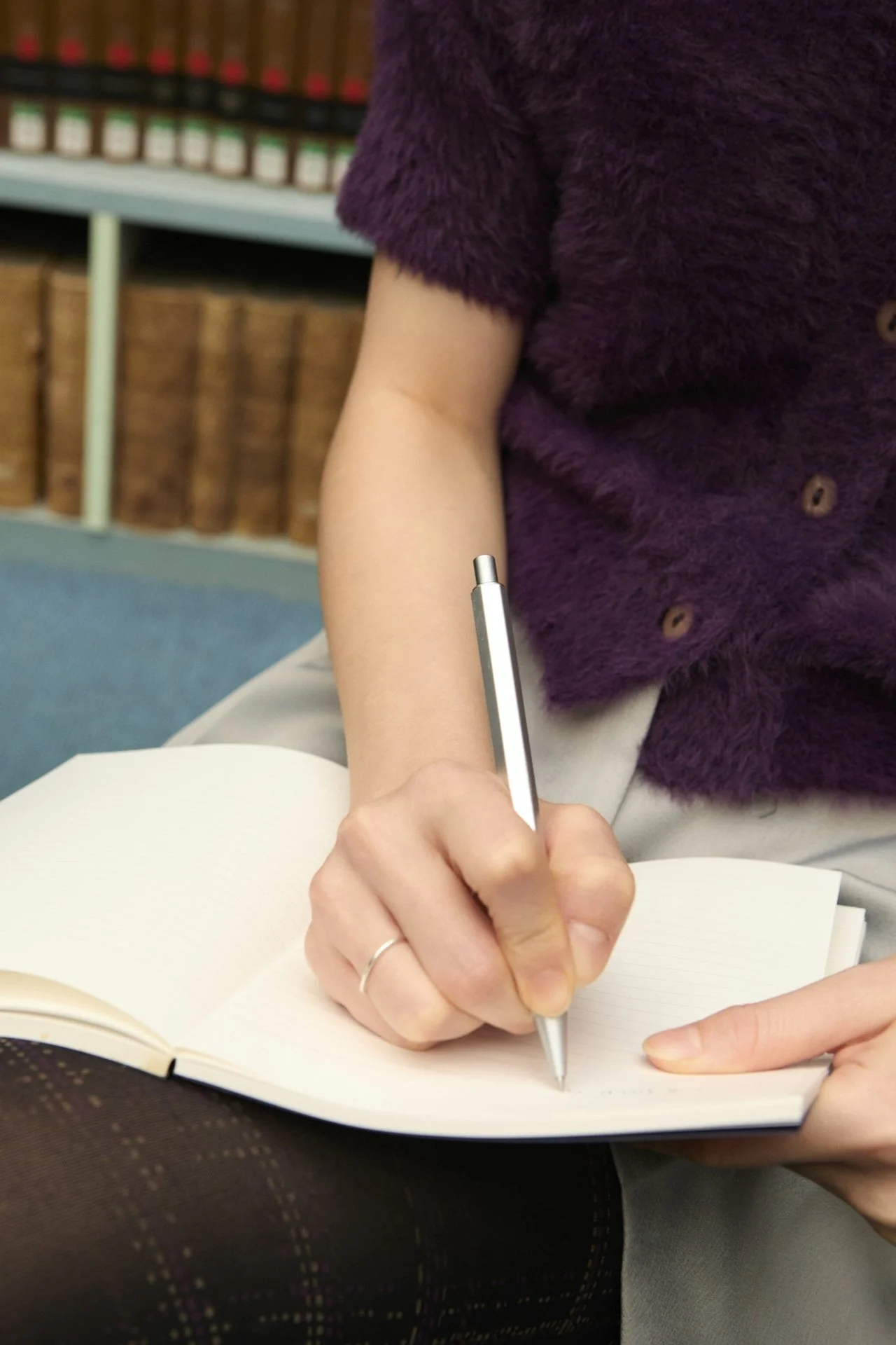 Person sitting with an open notebook and writing in it with a pen, wearing a purple fluffy sweater and measuring tights, books on shelves in the background.