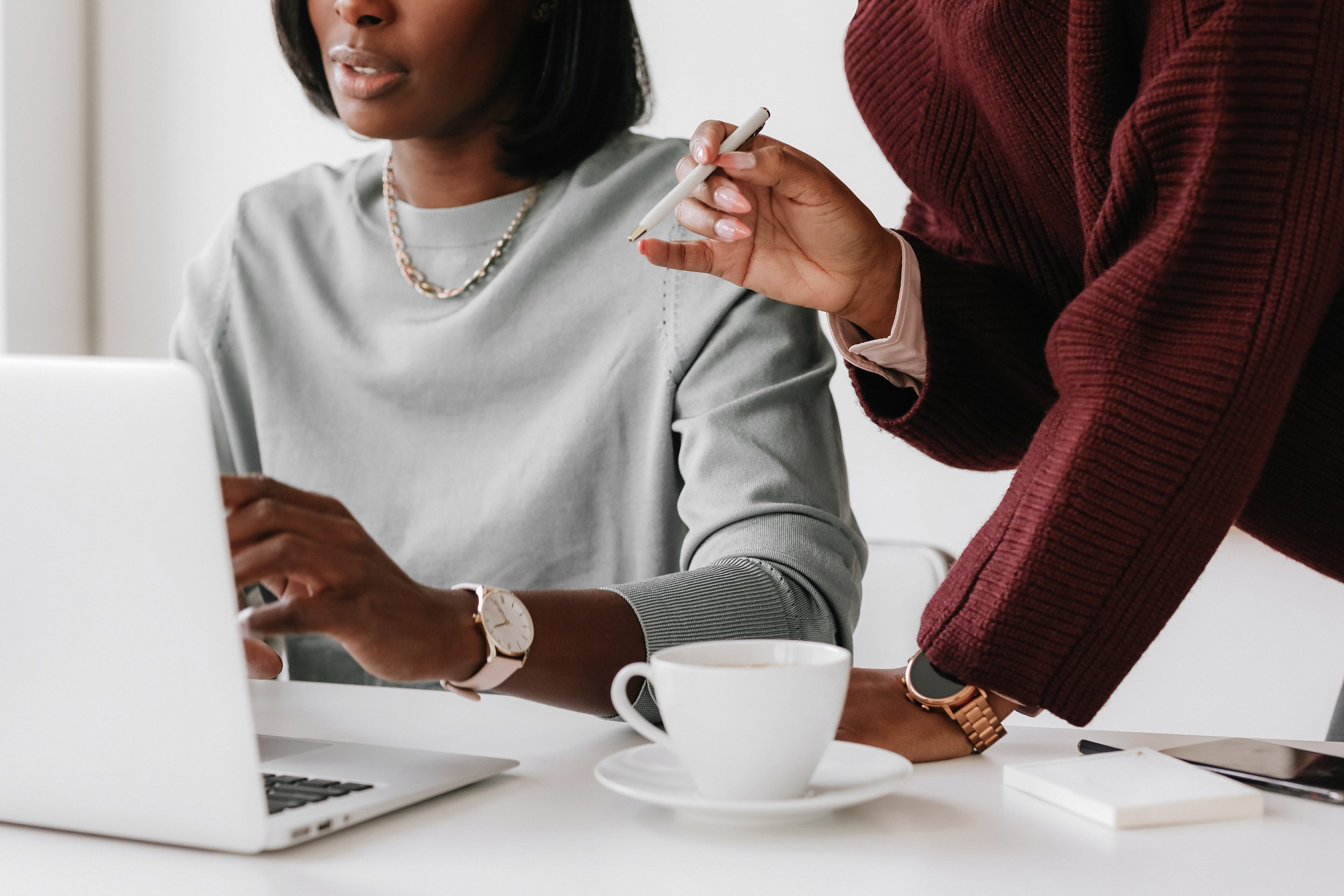 Two women working together at a desk, one using a laptop and the other holding a pen, with a coffee cup on the table.