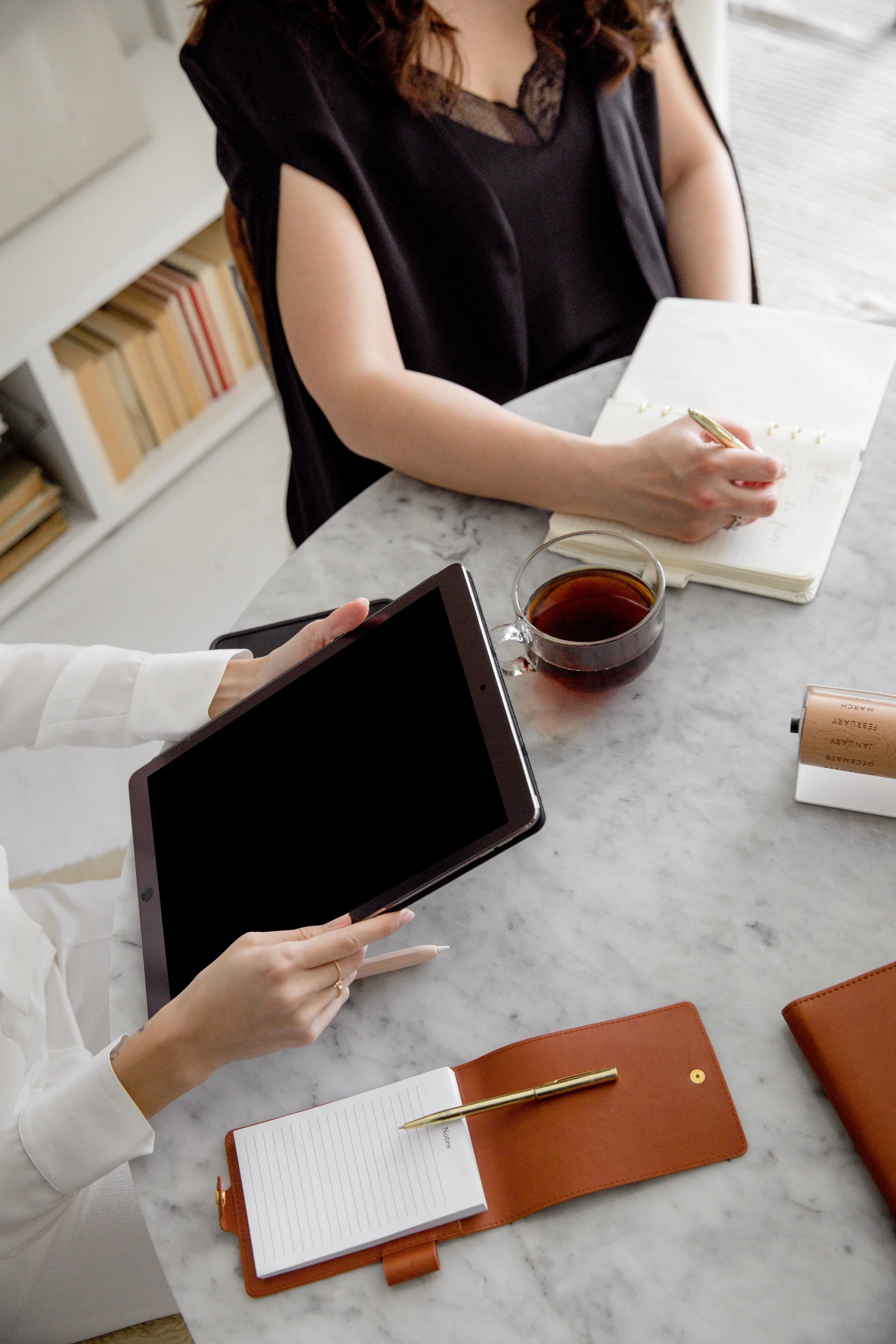 Two women sitting at a marble table, one holding a tablet and the other writing in a notebook. There is a cup of tea, a small notebook, and a brown leather wallet on the table.