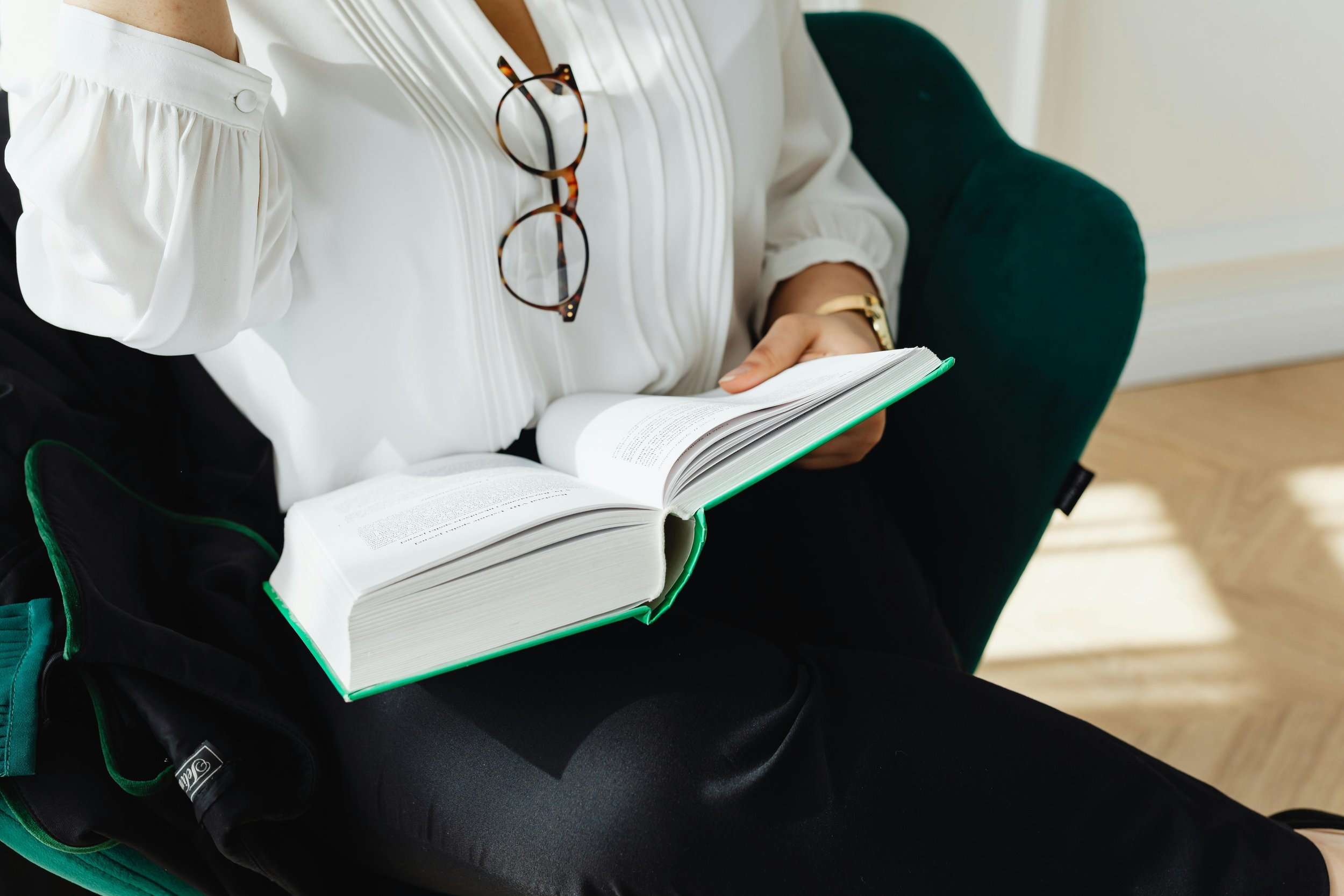 A woman sitting on a green chair, reading an open book while wearing glasses around her neck and a white blouse.