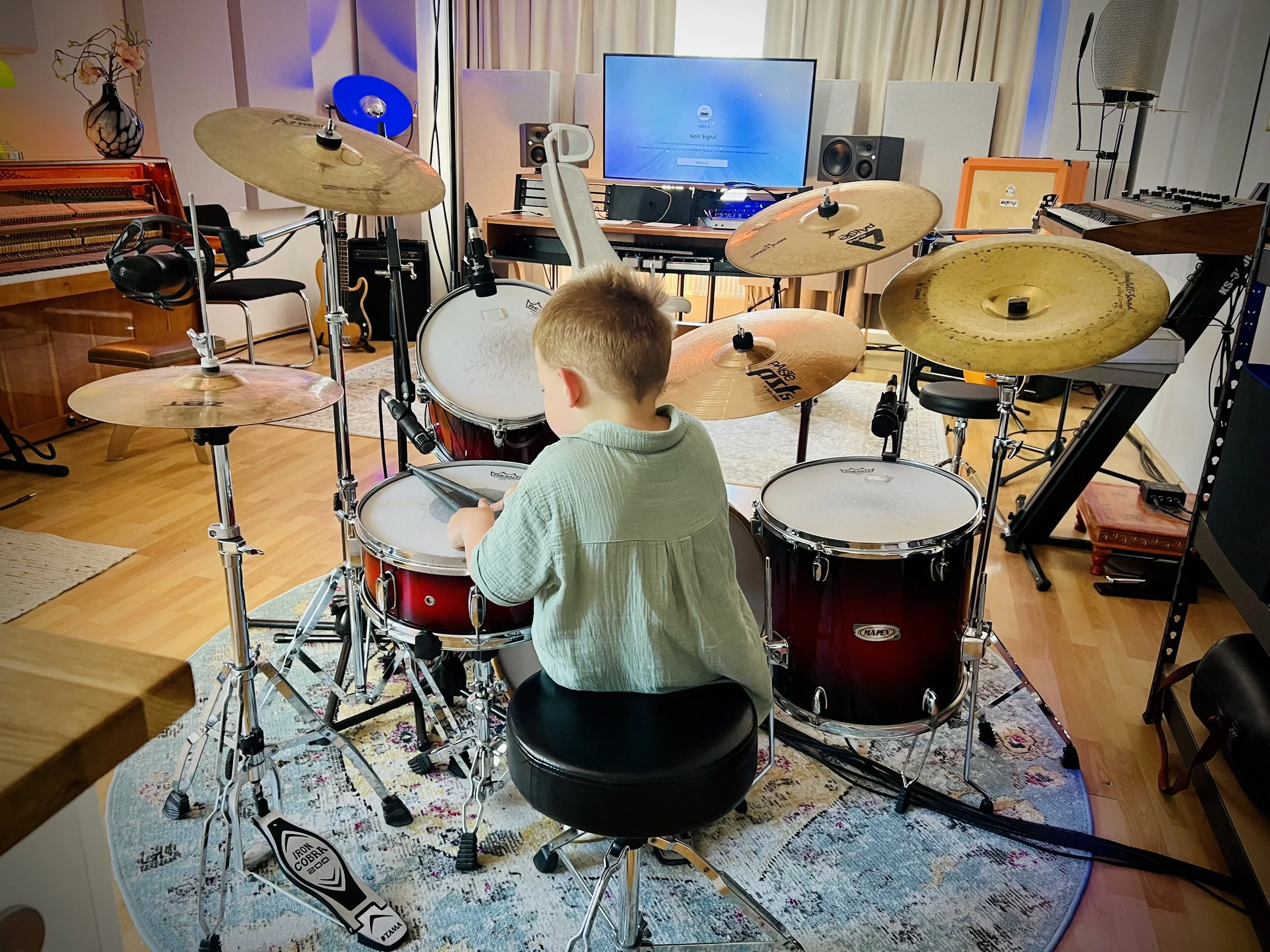 Ein junge spielt auf einem elektronischen Schlagzeug in einem Musikzimmer, umgeben von Musikinstrumenten und einem großen Fernseher.