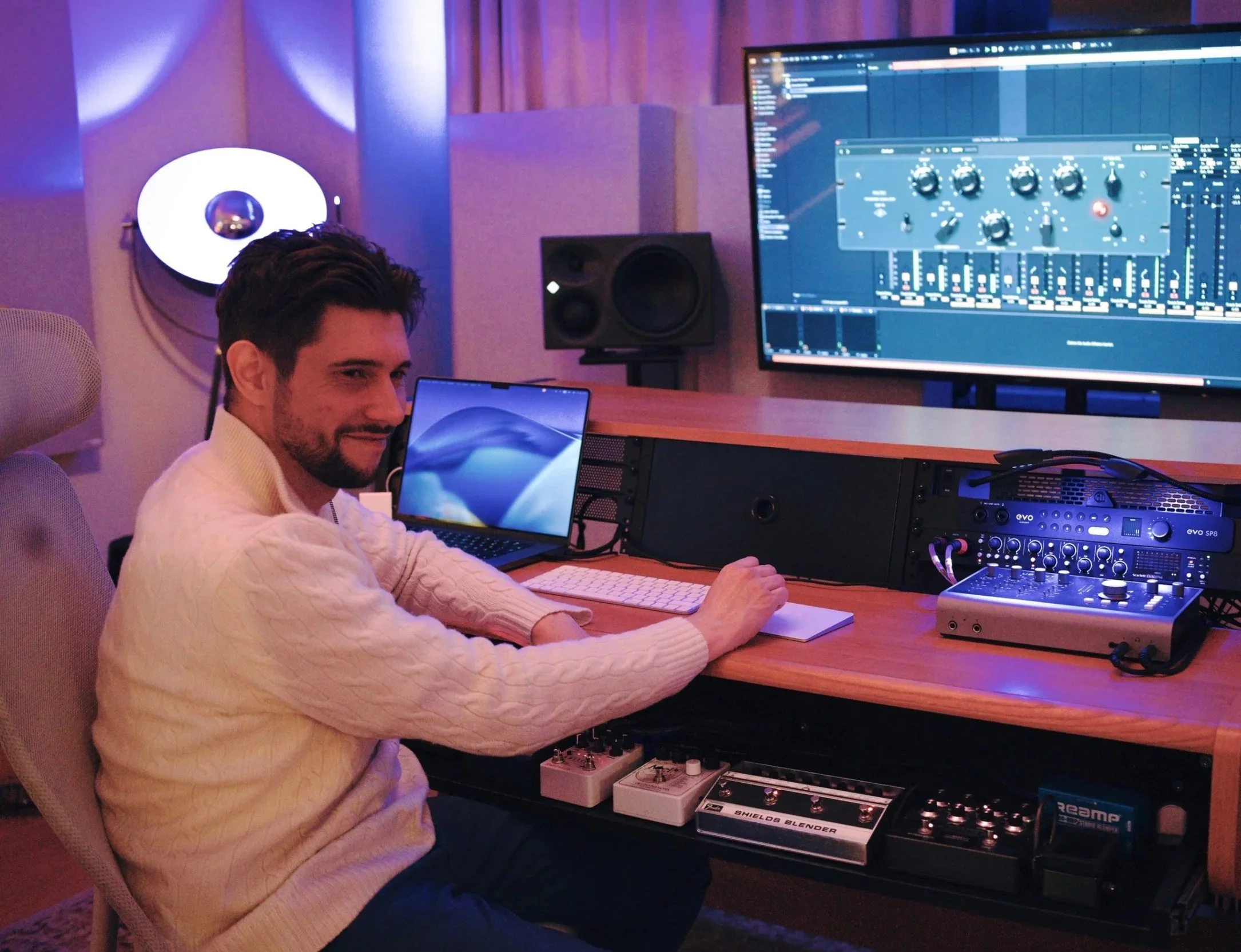 Ein Mann sitzt in einem Tonstudio vor einem Computerbildschirm mit Musiksoftware und Audio-Equipment, Lichter in Blau- und Lilatönen im Hintergrund.