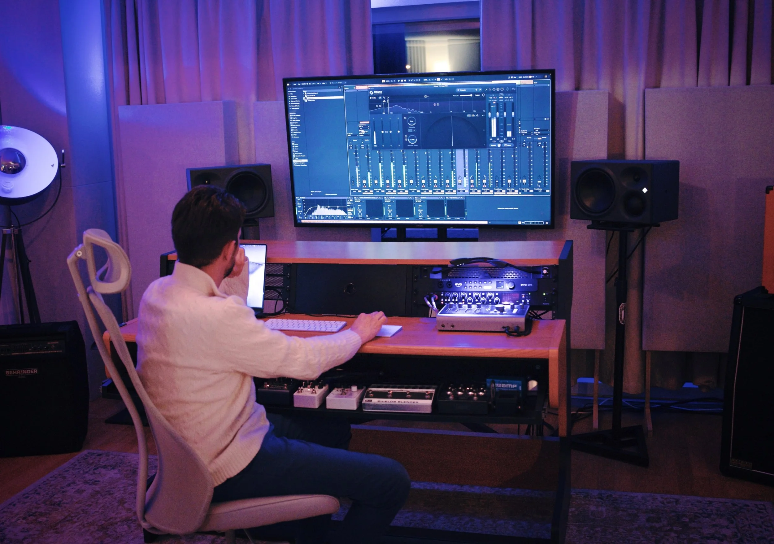 Ein Mann arbeitet in einem Tonstudio an einem Computer mit Musiksoftware. Der Raum ist gedämpft und mit akustischen Wandpaneelen ausgestattet, es sind Lautsprecher und Musikelektronik auf einem Tisch vor ihm installiert.
