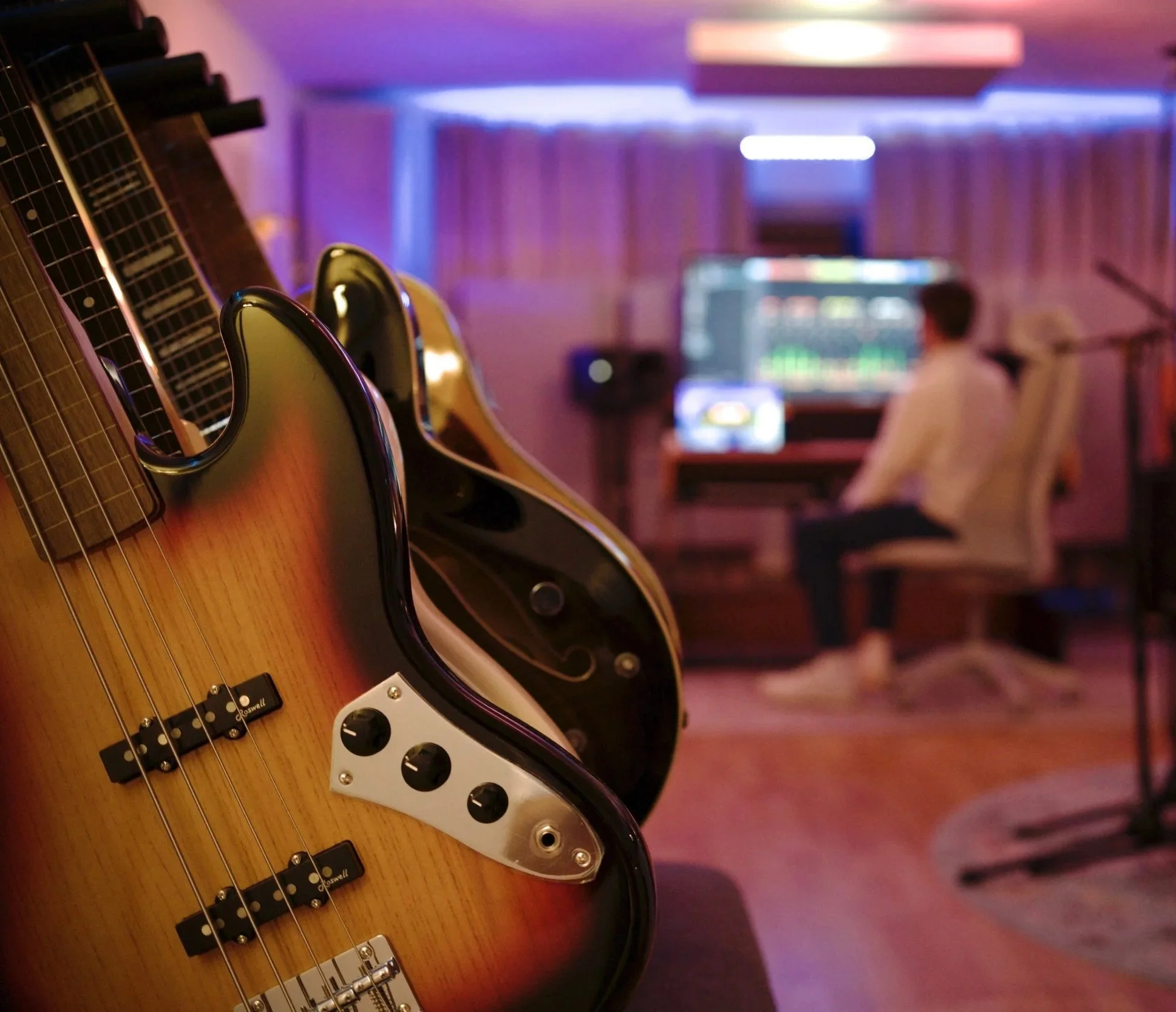 E-Gitarre liegt auf einem Tisch in einem Raum mit bunten Lichteffekten, im Hintergrund ist eine Person vor einem Bildschirm zu sehen.