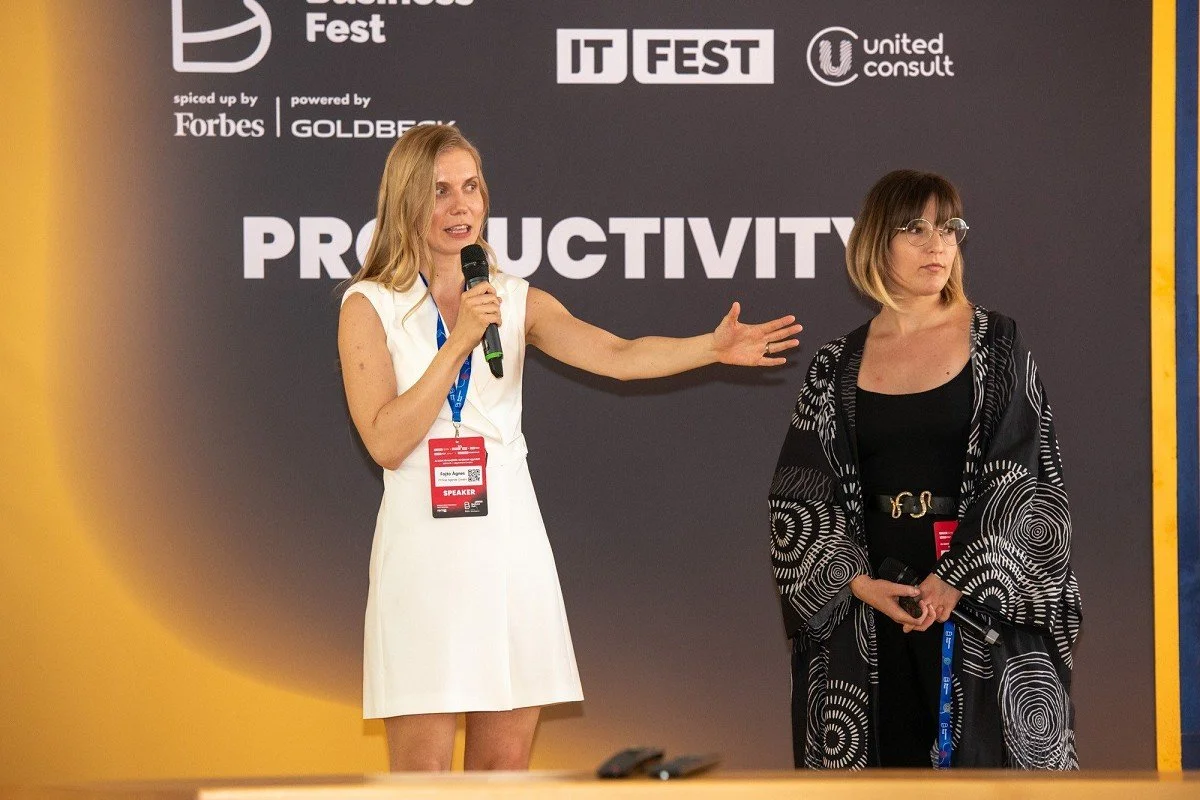 Two women speaking on stage at a productivity conference or event, with a dark background displaying sponsor logos and the word 'PRODUCTIVITY' in large letters. One woman is holding a microphone and gesturing, while the other woman is standing next to her, holding a microphone.