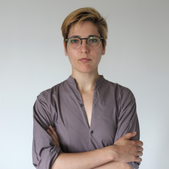 A woman with short light brown hair, glasses, and a gray shirt, standing with arms crossed against a plain white background.