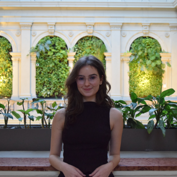 A young woman with glasses and long brown hair, wearing a sleeveless black top, standing in front of a decorative indoor garden with lush green plants and architectural arches.