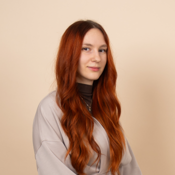 Young woman with long red hair wearing a gray sweater, standing against a beige background.