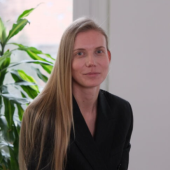 A woman with long blonde hair wearing a black blazer sitting indoors near a large green plant.