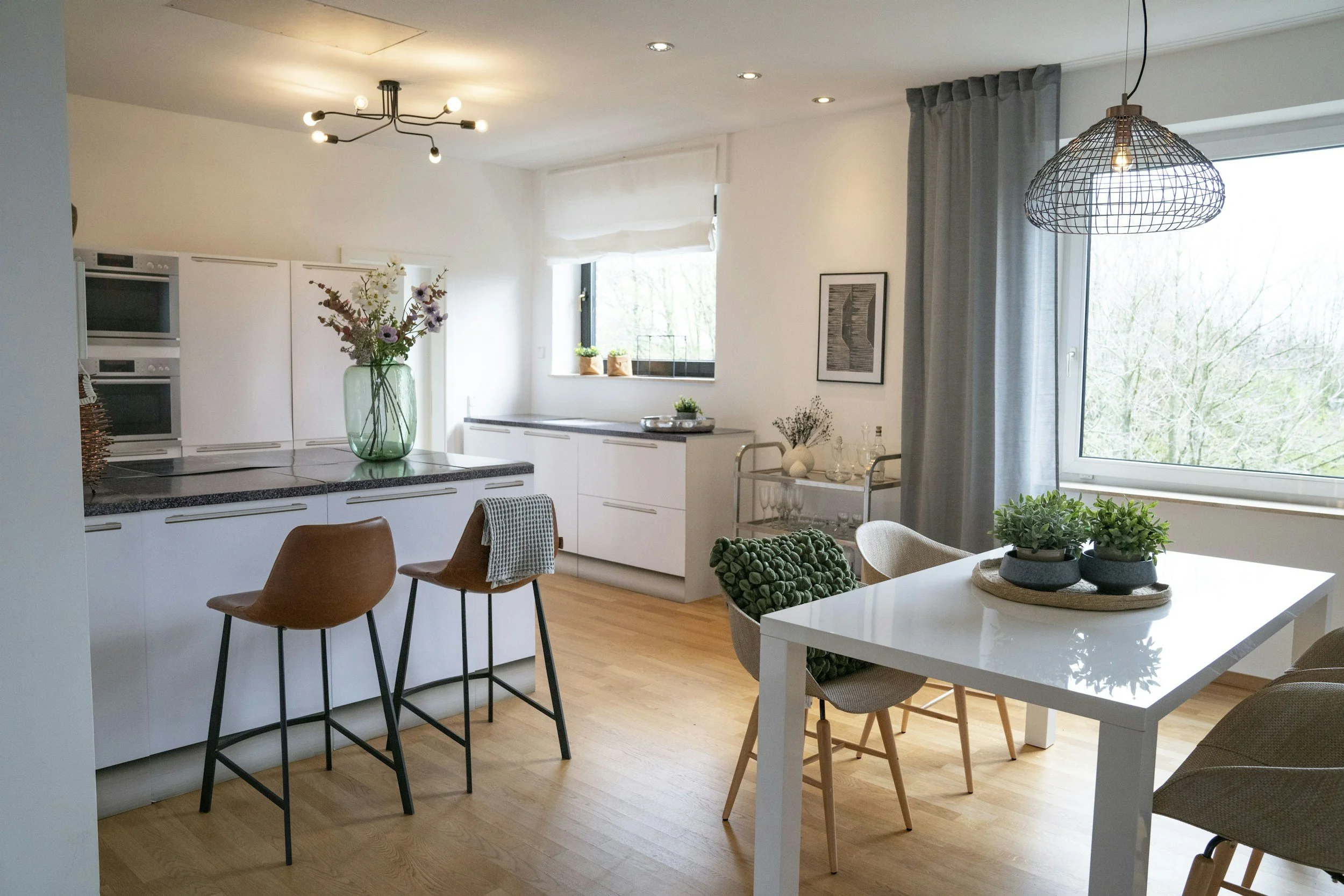 Modern open-plan kitchen and dining area with white cabinets, a window, and wooden flooring, decorated with green plants and minimalist lighting fixtures.