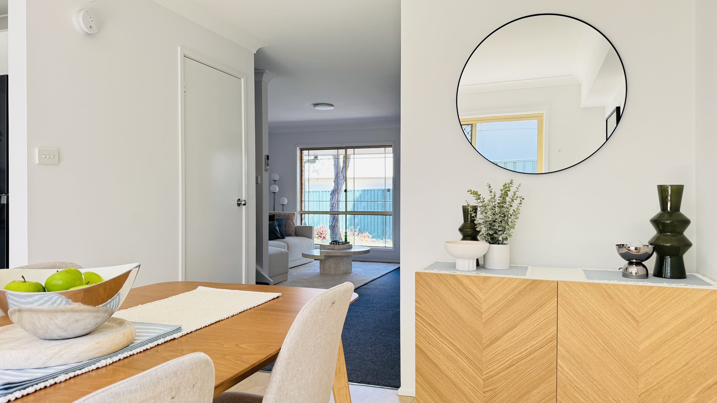 Interior view of a modern home, showing a dining area with a wooden table and beige chairs, a sideboard with decorative vases and a round mirror on the wall, and a living room visible through an open doorway with a window overlooking a backyard.