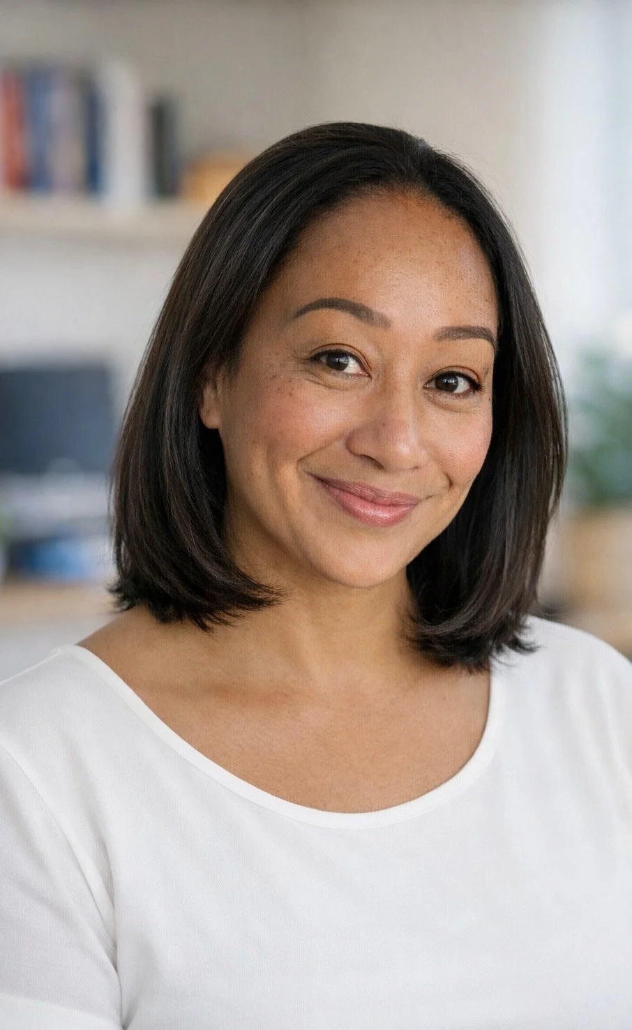 A woman with shoulder-length black hair, wearing a white top, smiling at the camera in a cozy indoor setting.