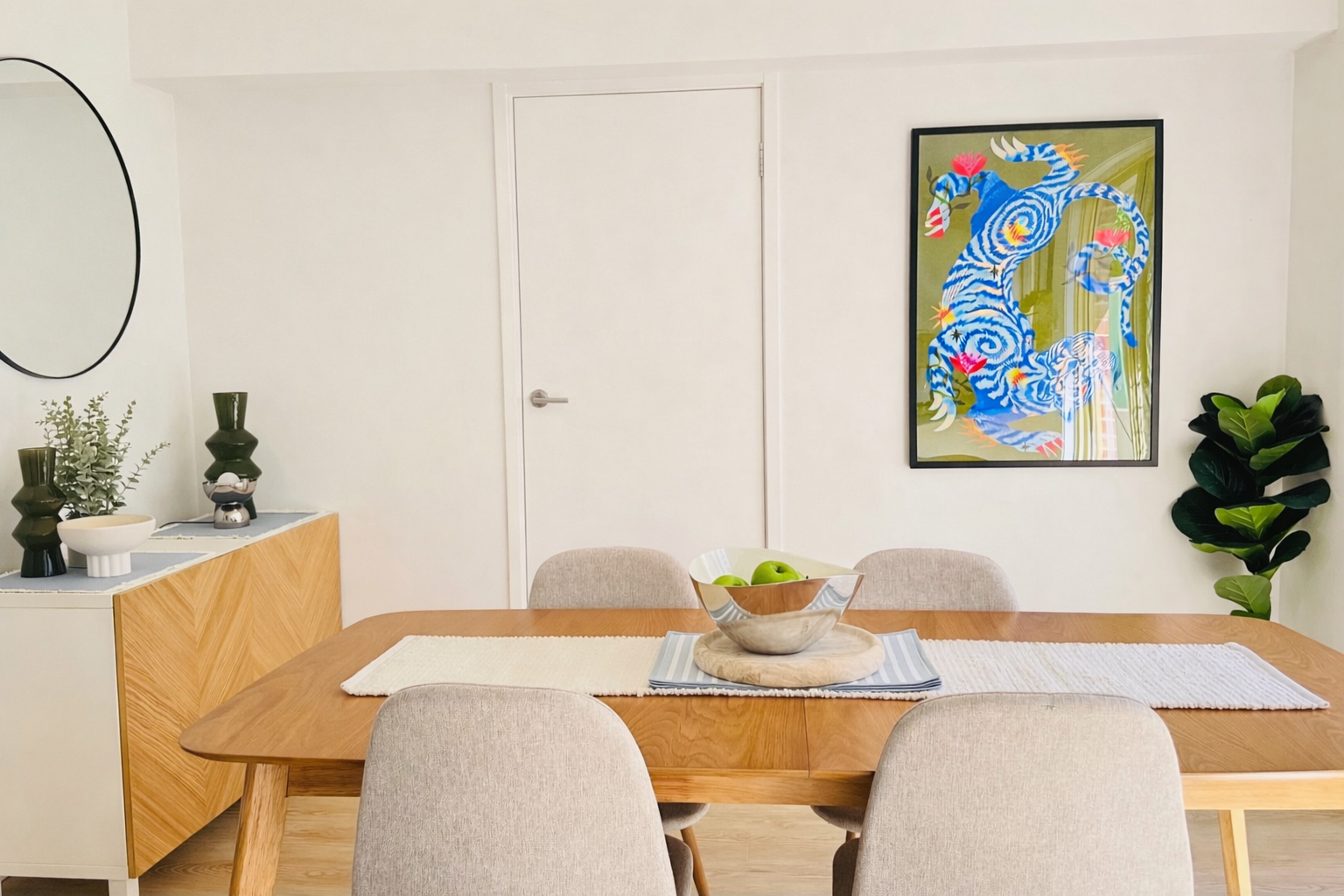Dining room with wooden table, four beige upholstered chairs, a white and gray striped table runner, a glass bowl of green apples, wall art featuring a colorful blue and green abstract design, a sideboard with decor items including vases and plants, and a large green leafy plant in the corner.