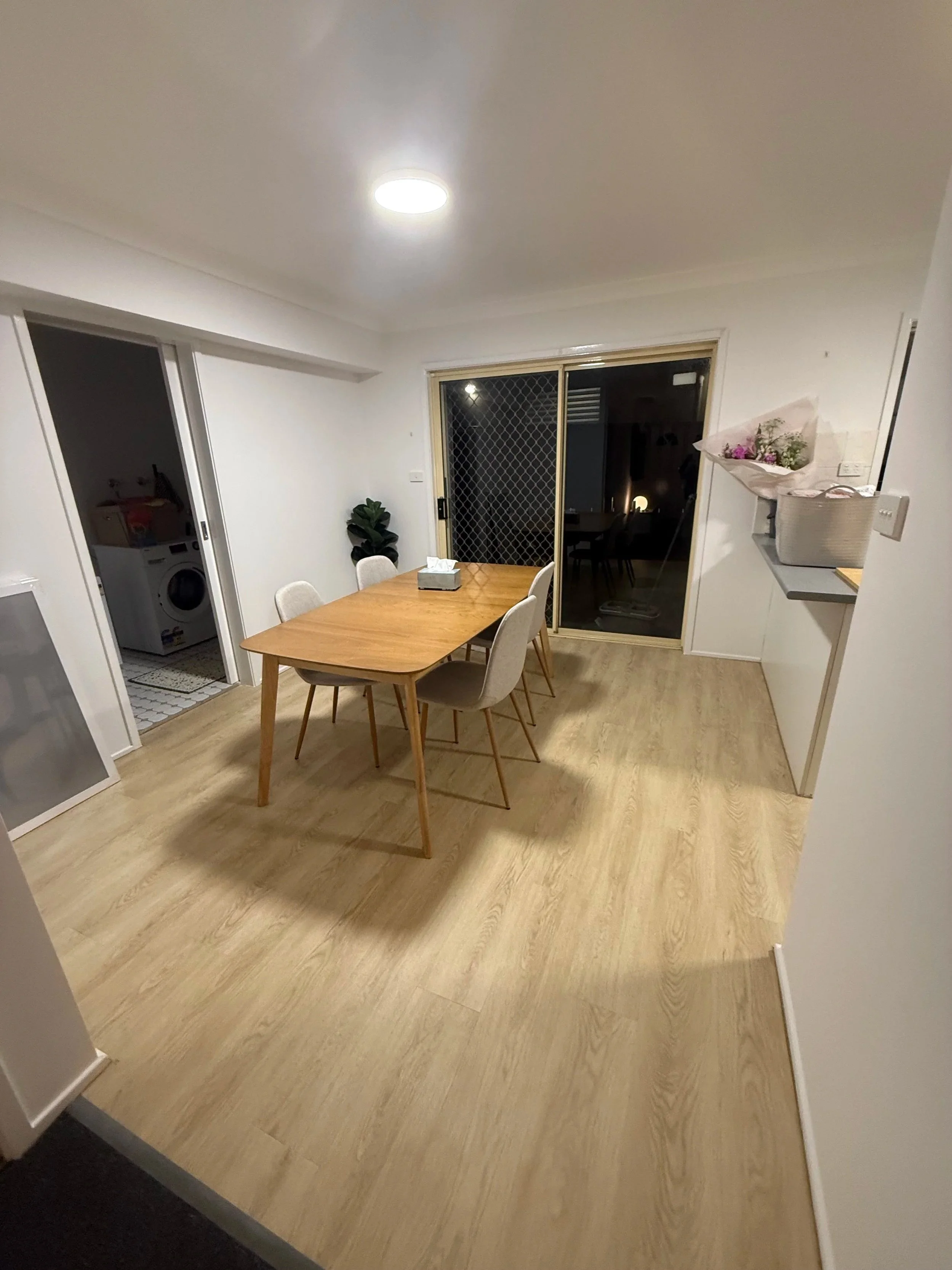 A dining area with a wooden table, light-colored chairs, sliding glass door, and a laundry room visible through an open door with a washing machine inside.