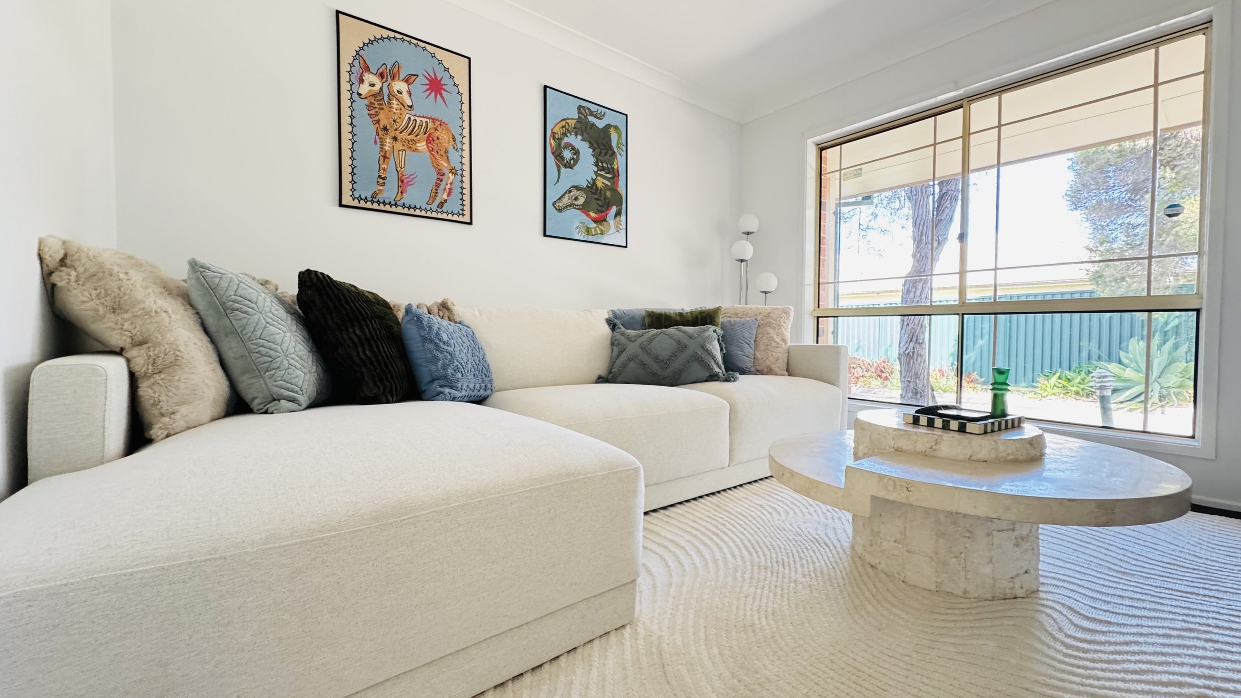 A bright living room with a white sectional sofa decorated with multiple textured and colored throw pillows. Modern artwork hangs on the wall, and a large window lets in natural light. A round stone coffee table is positioned in front of the sofa, holding a striped tray, a small green vase, and a green decorative candle. A tall floor lamp with multiple white globes stands near the window.
