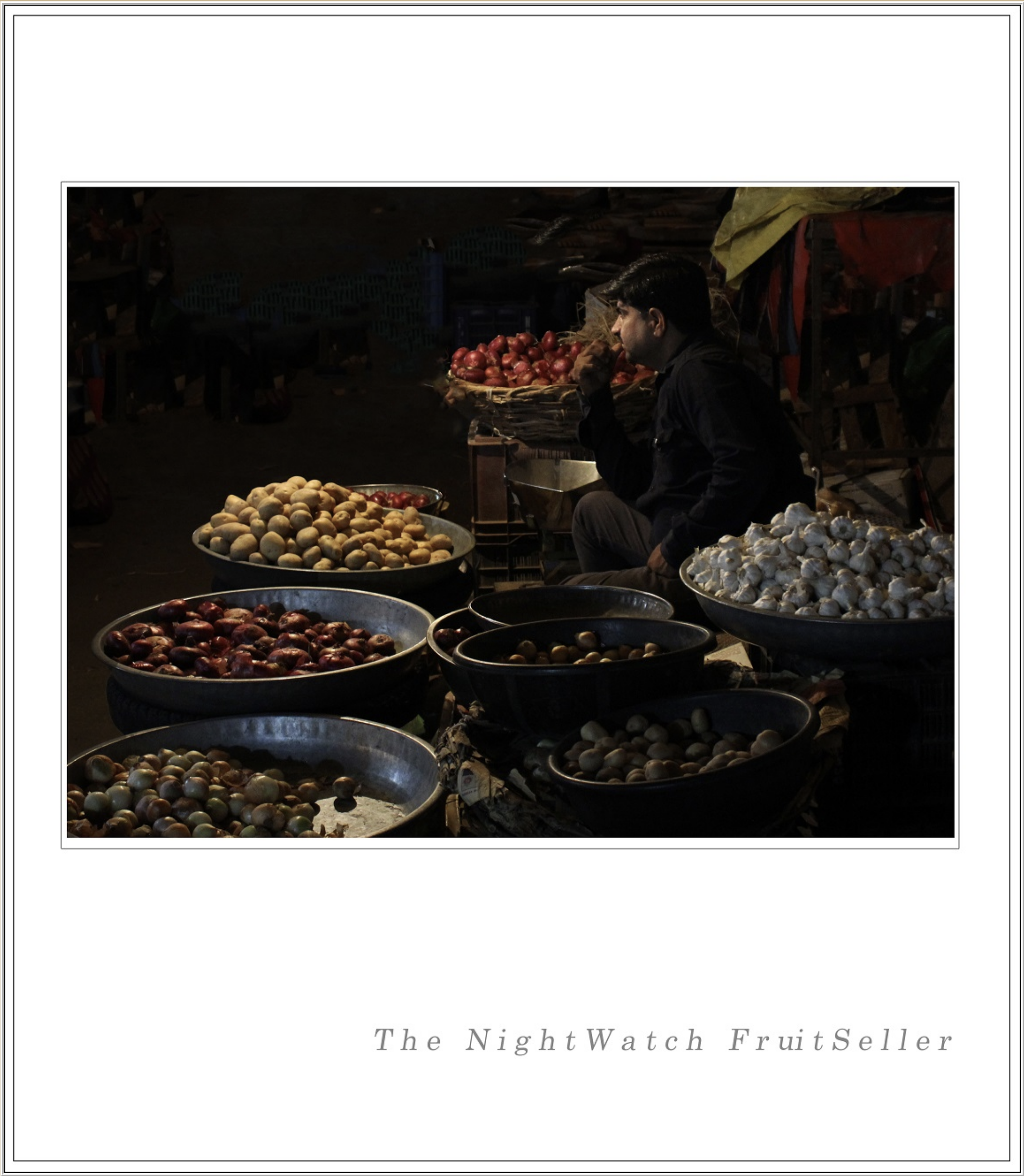 Night scene of a market stall displaying piles of potatoes, onions, and garlic with a person sitting behind the stall in profile.
