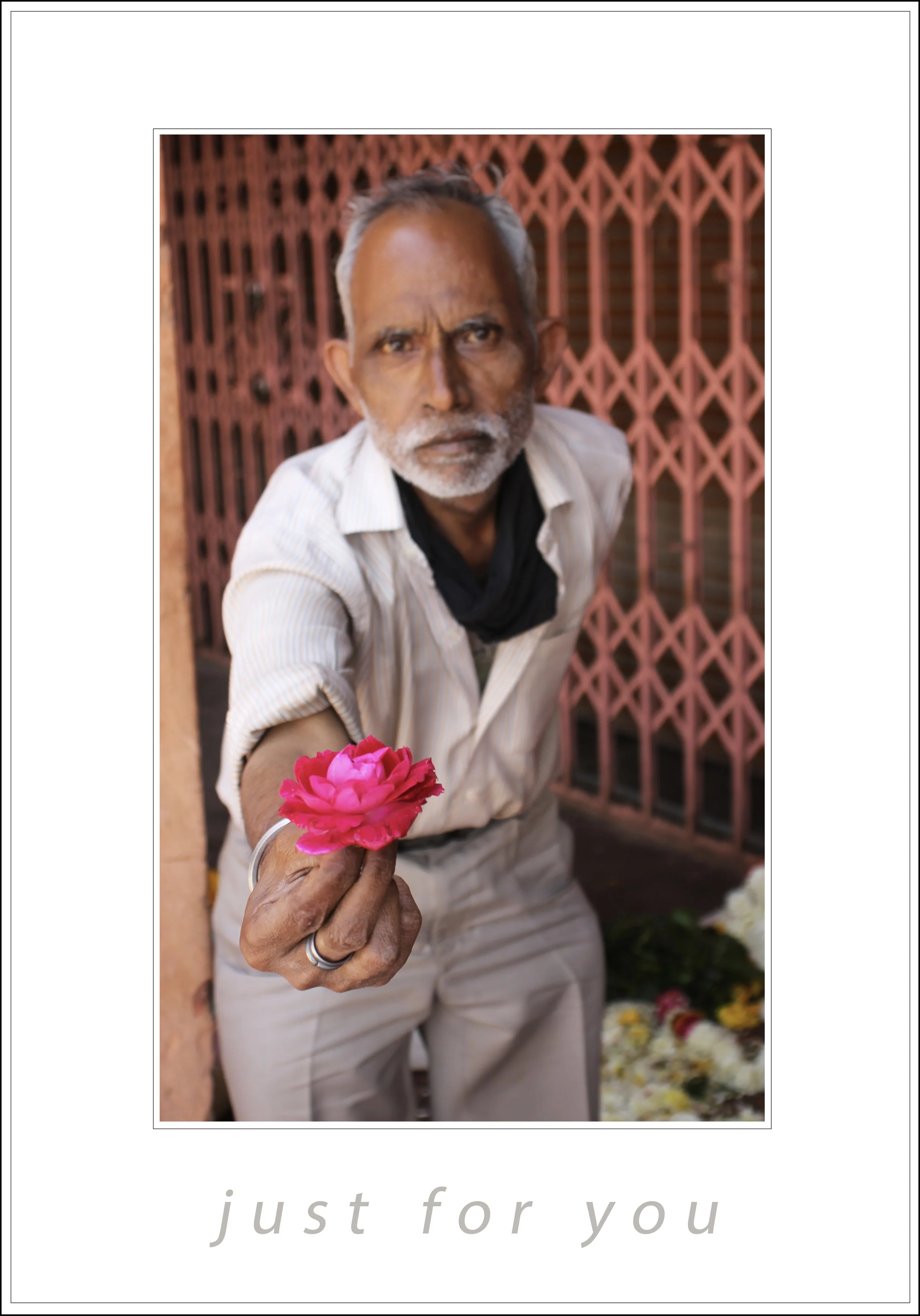 An older man with gray hair and beard offering a pink flower, standing in front of a decorative red iron gate, with soft lighting highlighting his serious expression.
