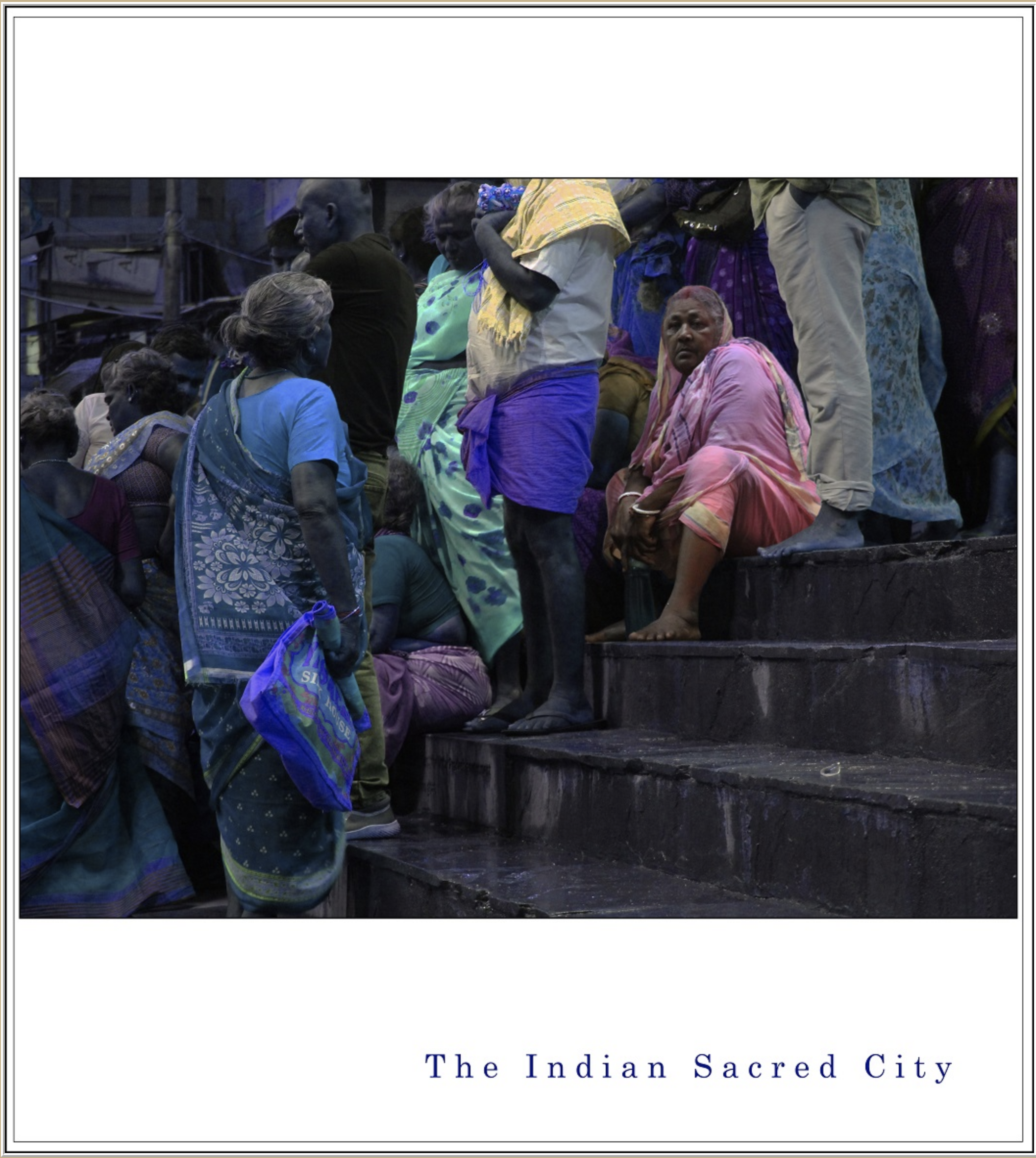 A group of Indian women and men sitting and standing on steps, wearing traditional colorful sarees and other clothing, engaged in a gathering or ceremony, with a backdrop of buildings and electrical wires.