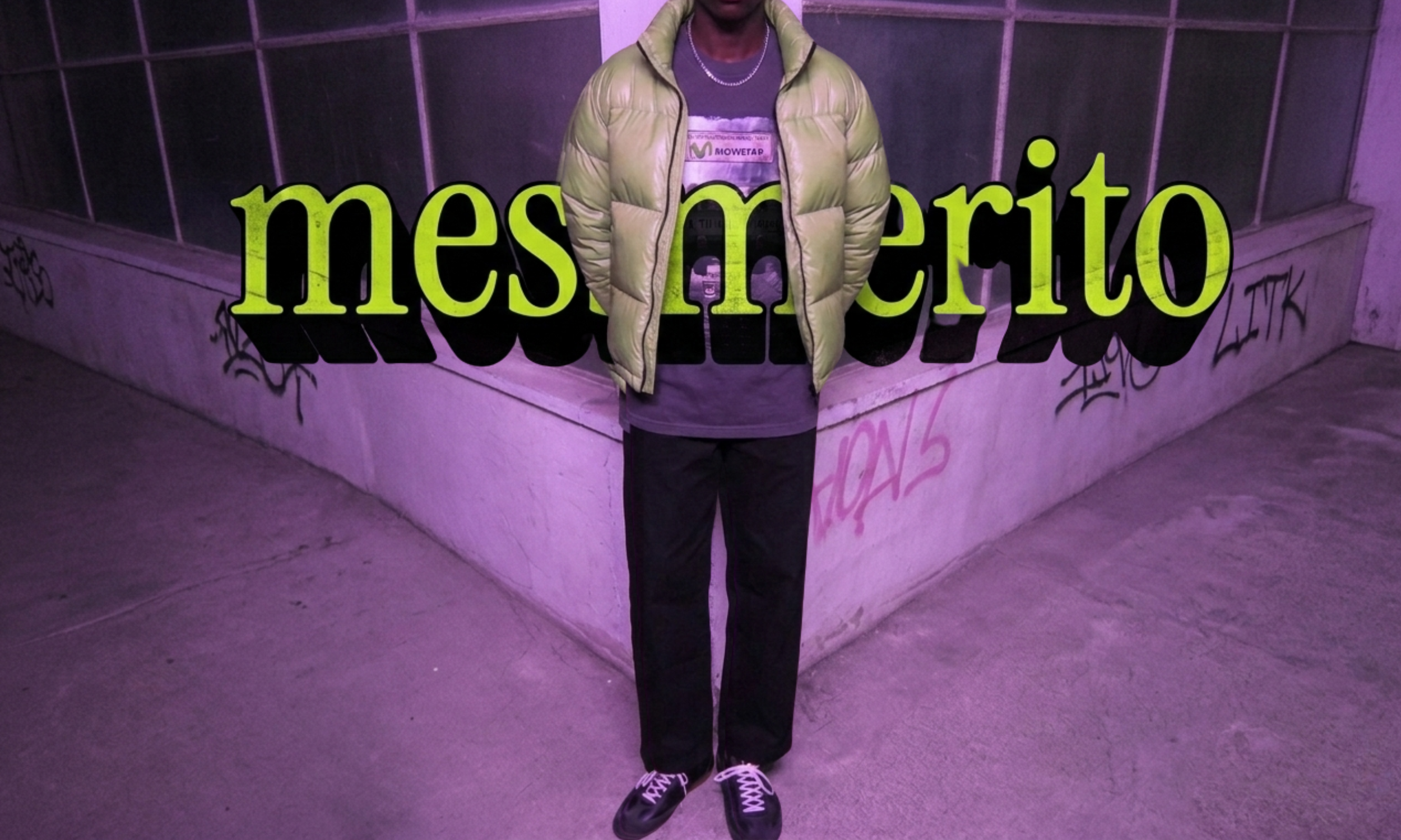 A person standing in front of a Graffiti-covered, pink-lit concrete wall. The individual is wearing black pants, a gray shirt, a light green puffer jacket, black sneakers with white laces, and a silver chain necklace. There is large, green, stylized text overlayed on the image that says "mes merito."
