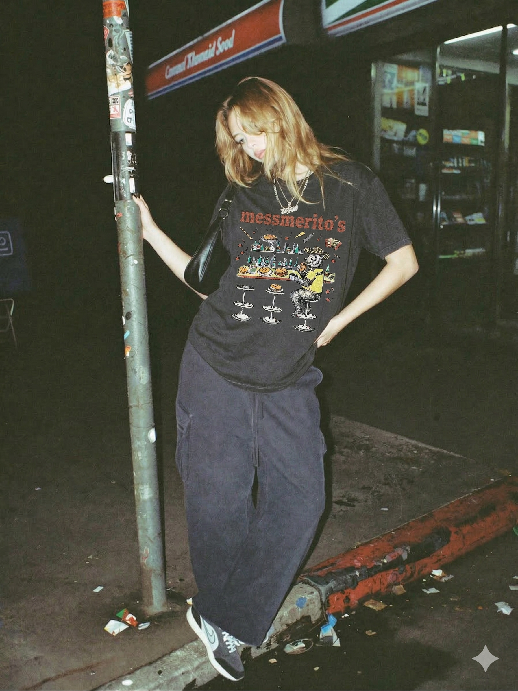 A woman with red hair standing outdoors at night, leaning against a street pole. She is wearing a black t-shirt with a graphic and the word 'messmerito's', dark pants, and sneakers. She has a black purse over her shoulder and is posing with a playful expression.