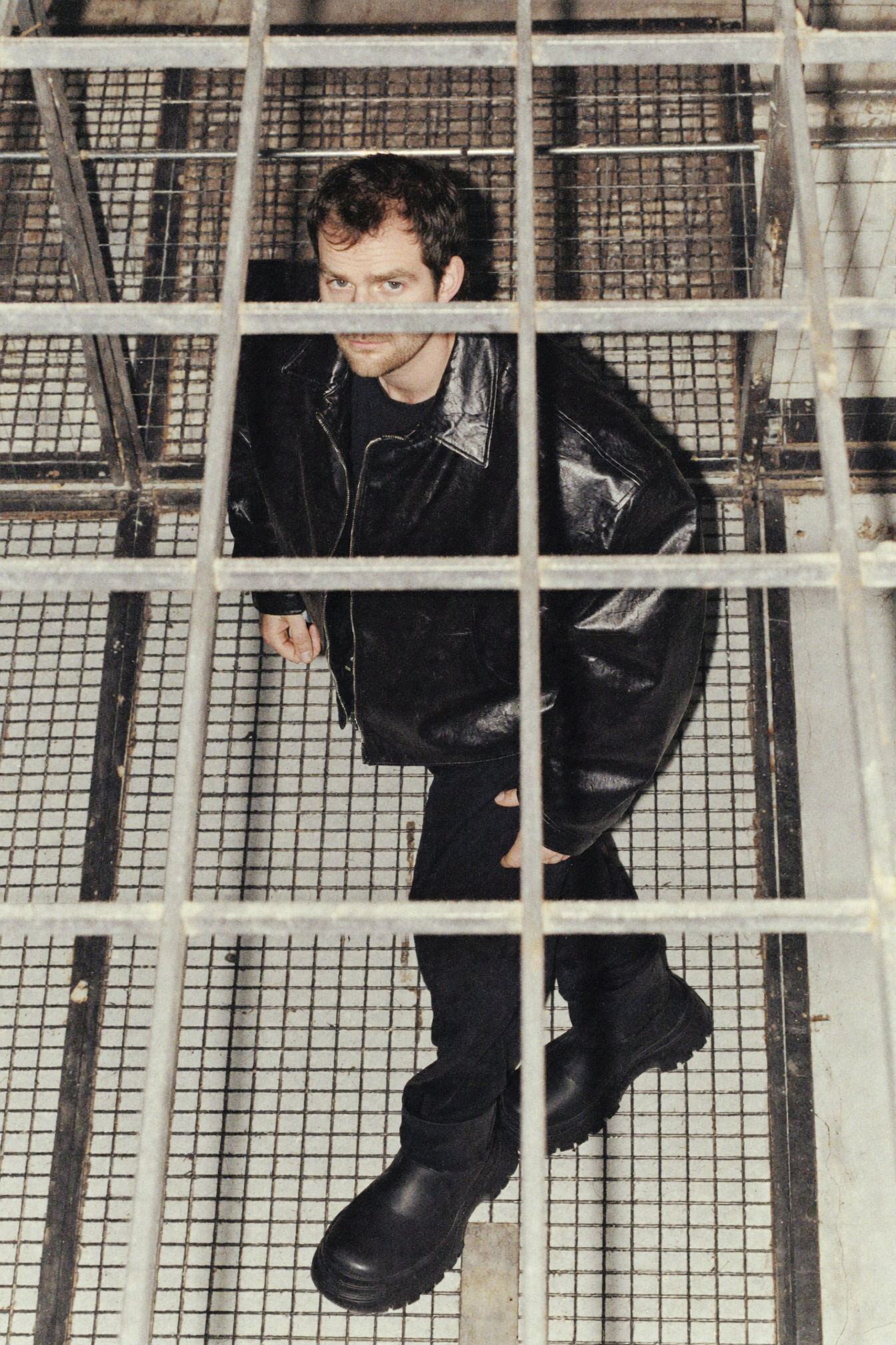 A young man with dark hair and a stubbled beard wearing a black leather jacket and black boots, bending down and looking up through a grid-like metal barrier in what appears to be an industrial or basement setting.