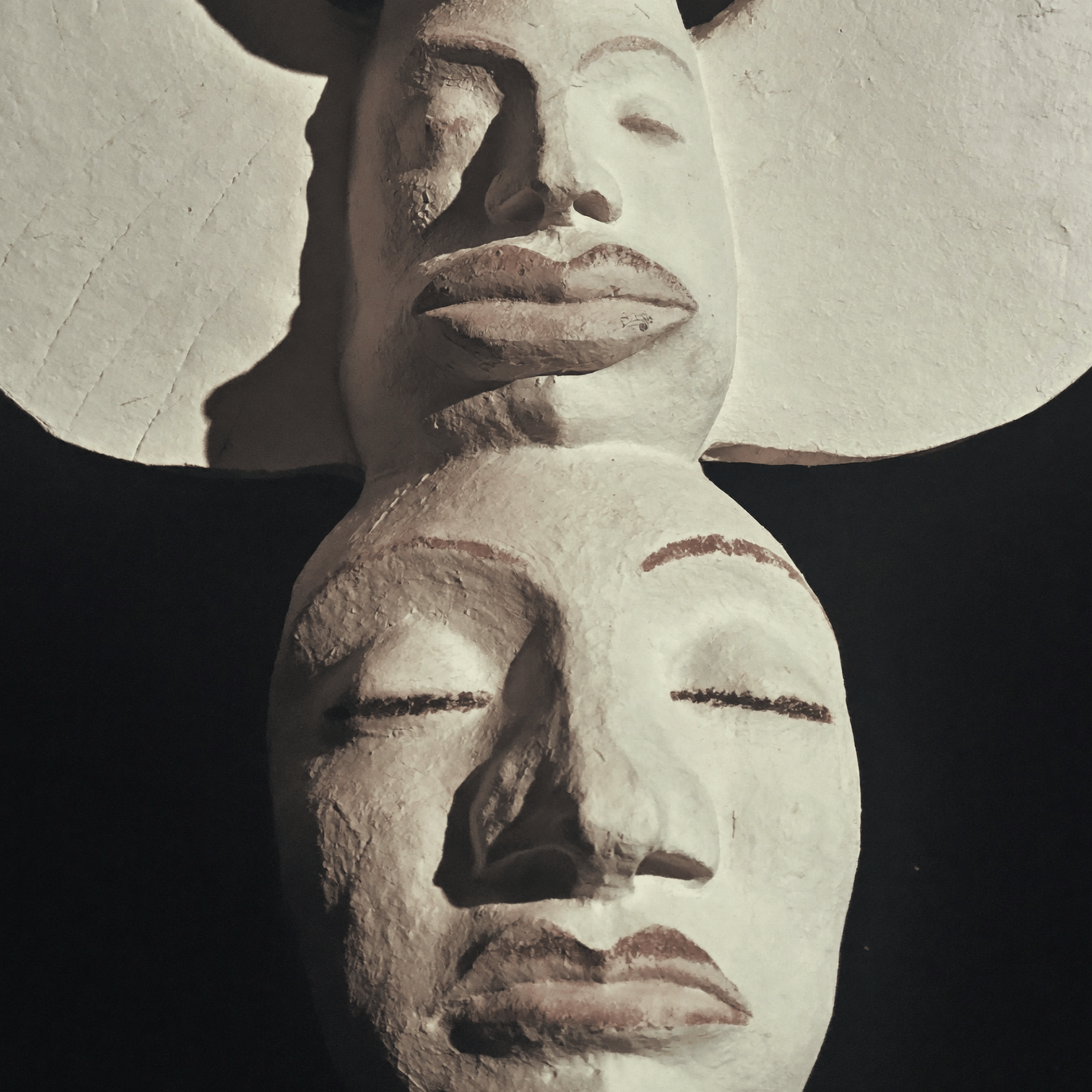 A textured sculpture of a human face with closed eyes, a mustache, and lips, mounted on a wall with soft lighting casting shadows.