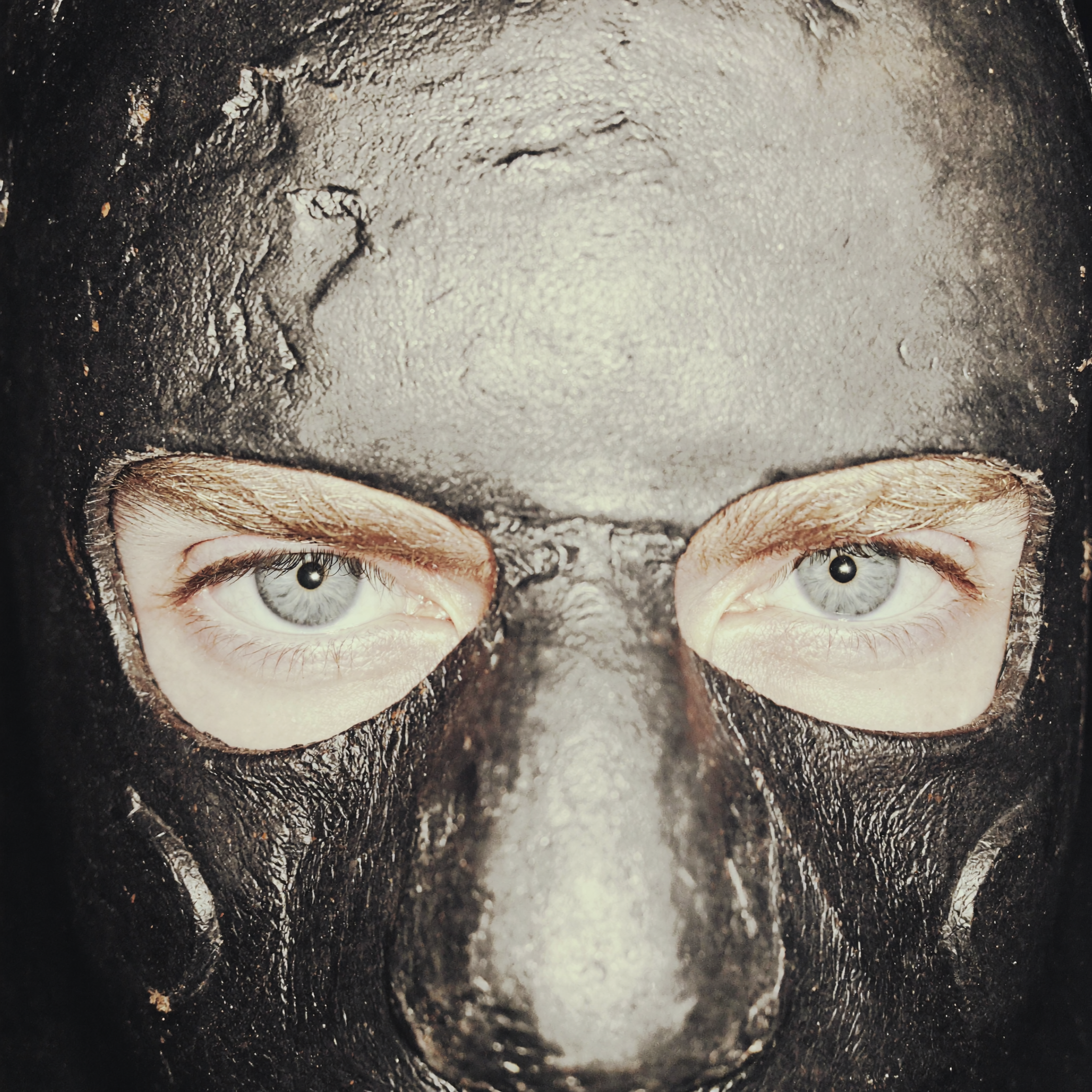 Close-up of a person's eyes and upper face covered in a dark, textured substance, possibly mud or clay, with the person's blue eyes peering through.