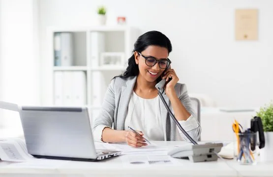 Woman sitting at a desk talking on the phone with a pen in hand, in a bright office with a laptop and office supplies.