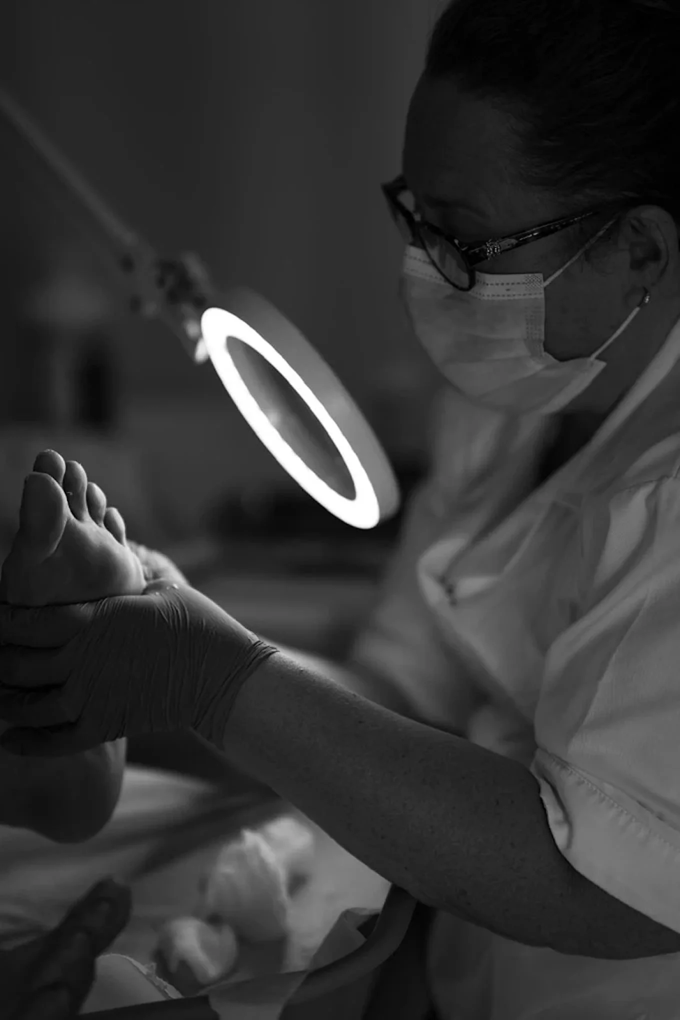 A woman wearing a surgical mask and gloves looks at the bottom of a foot with a light shining onto it