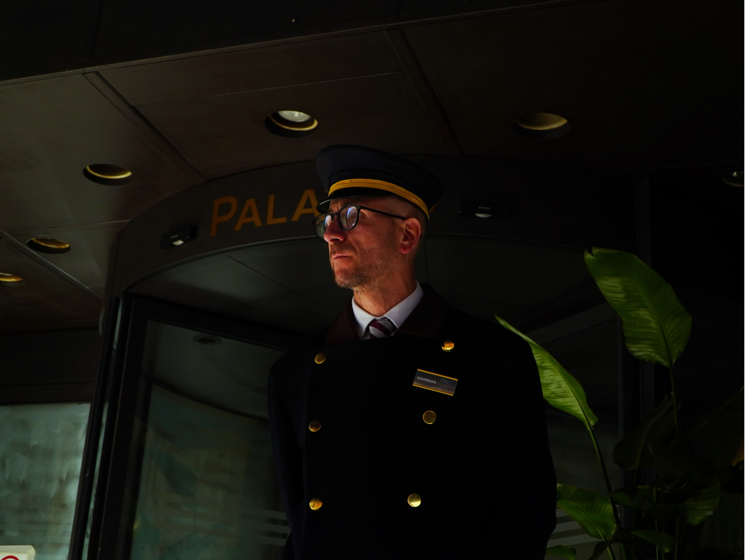Un gardien d'hôtel en uniforme noir, portant un chapeau et des lunettes, se tient dans un hall sombre avec quelques plantes vertes à sa droite, devant une entrée portant le mot "PALACE". Il a une expression sérieuse.