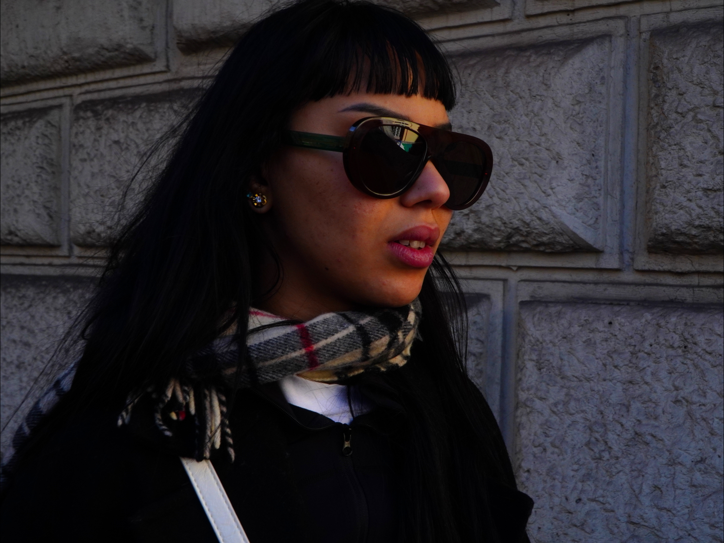 Jeune femme portant de grandes lunettes de soleil, un foulard à carreaux, un pull noir et un fond de mur en pierre