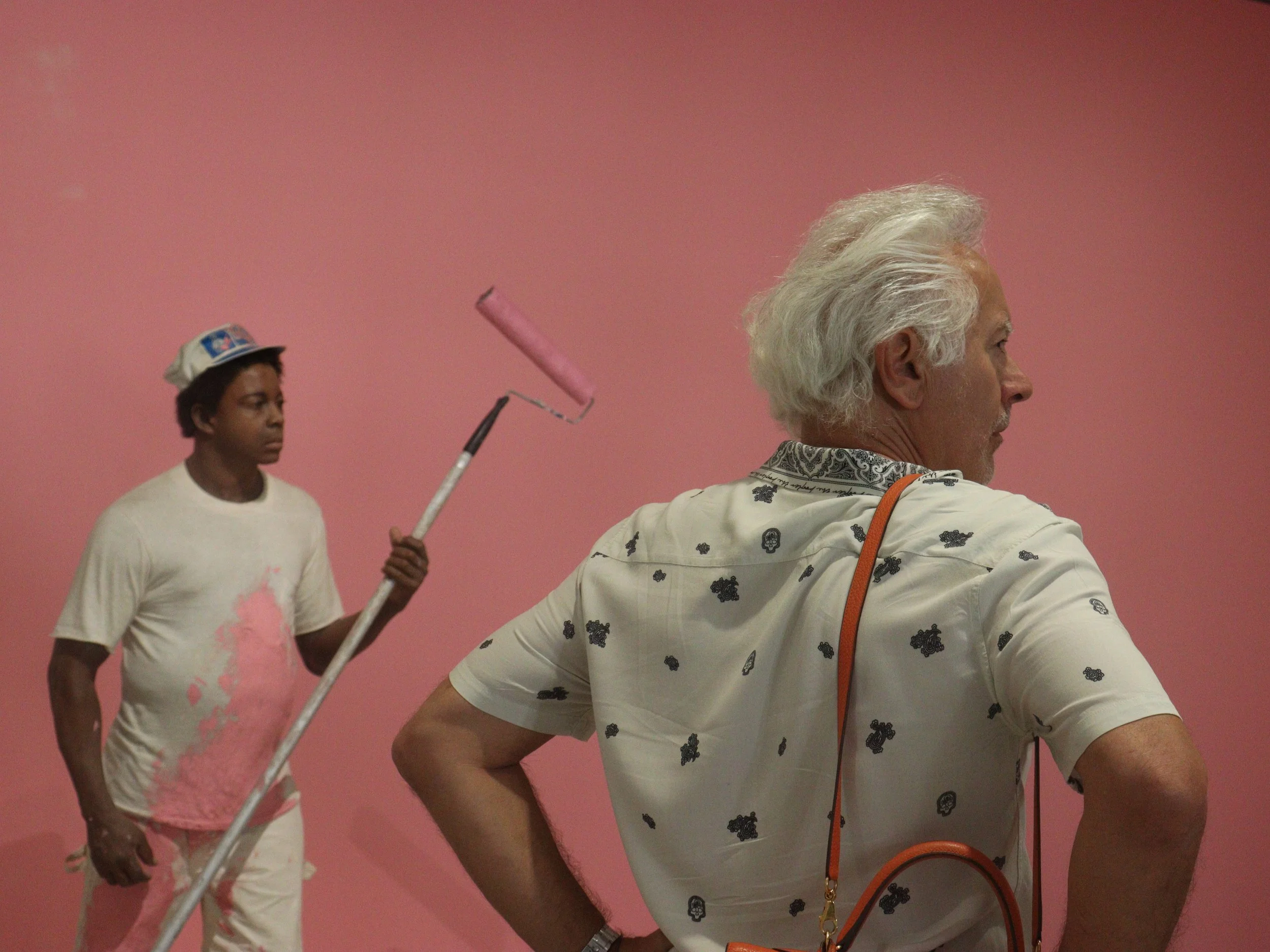 Un homme âgé avec des cheveux blancs regarde une œuvre d'art, un jeune homme noir avec un chapeau et un t-shirt blanc peint en rose. L'arrière-plan est rose