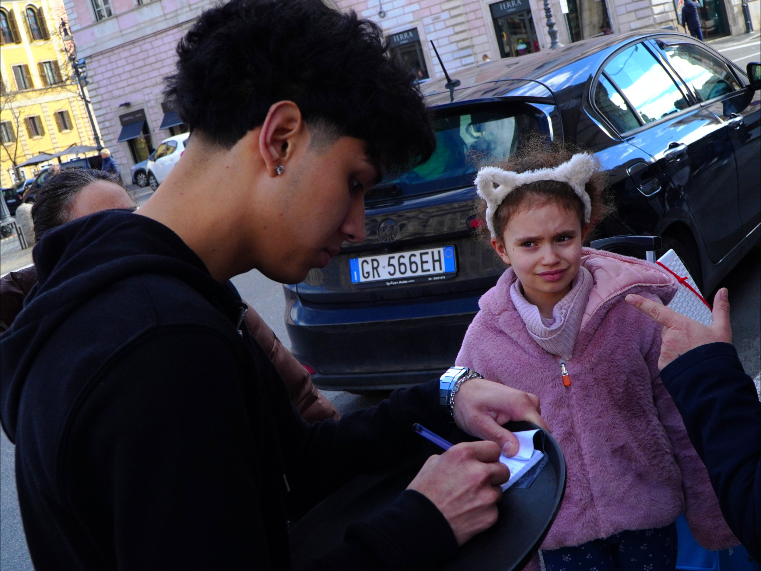 Jeune fille avec une écharpe rose et un bonnet en forme d'oreilles d'ours, qui semble confus ou contrariée, face à une jeune personne qui écrit dans un carnet, dans une rue urbaine.