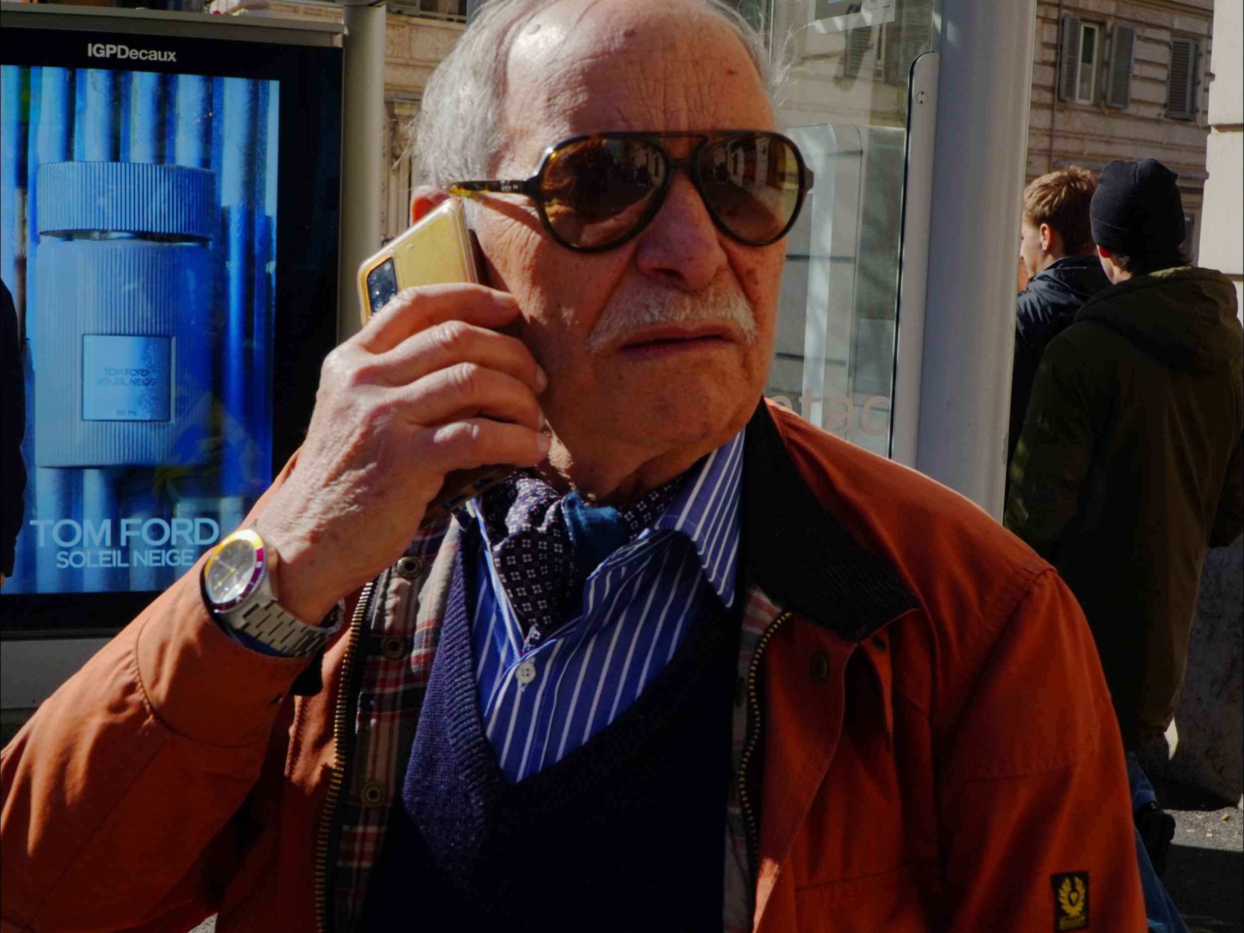 Un homme âgé portant des lunettes de soleil, une chemise à rayures, un pull et une veste orange, parle au téléphone dans une rue animée.