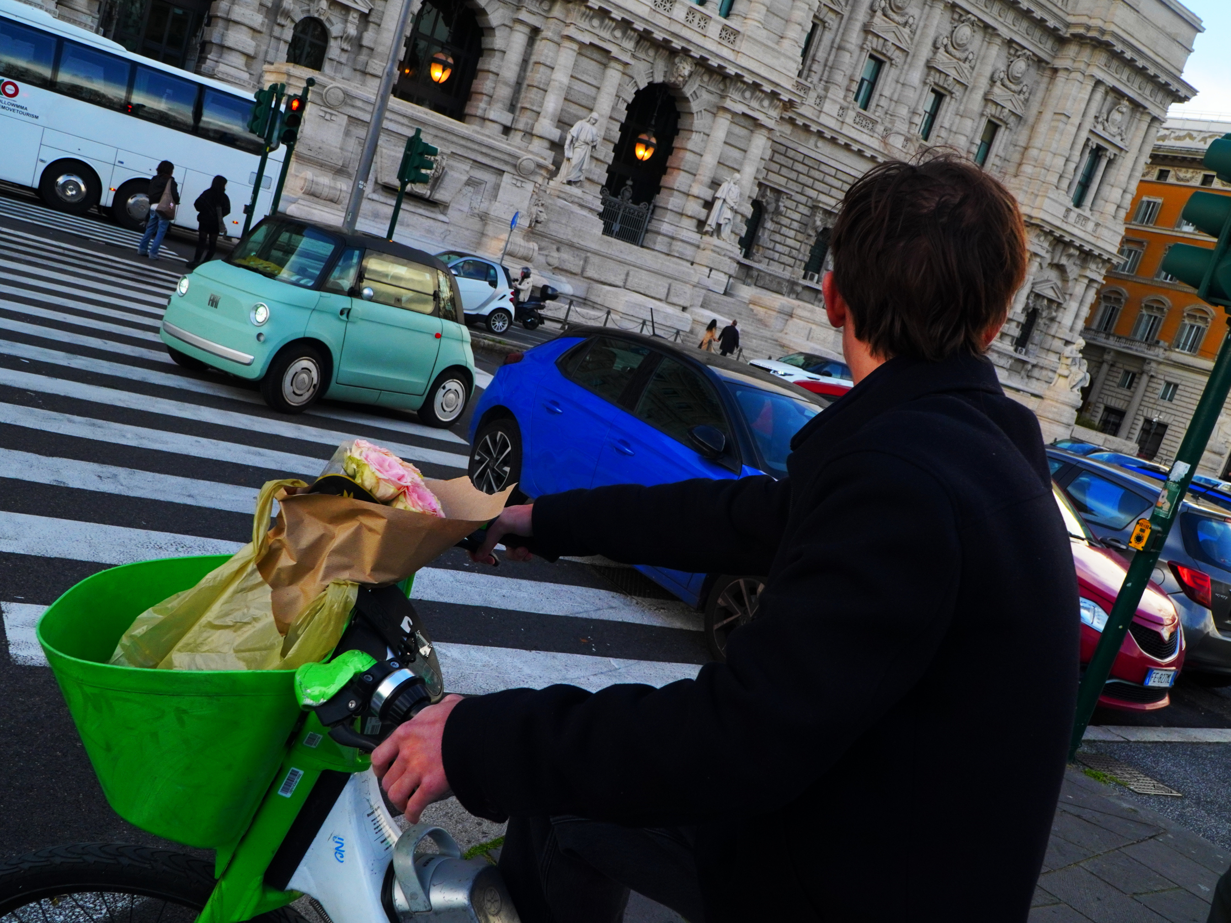 Une personne à vélo dépose un bouquet de fleurs dans un panier sur le vélo, en face d'un bâtiment historique en pierre, avec des véhicules garés sur la rue et des piétons en arrière-plan.