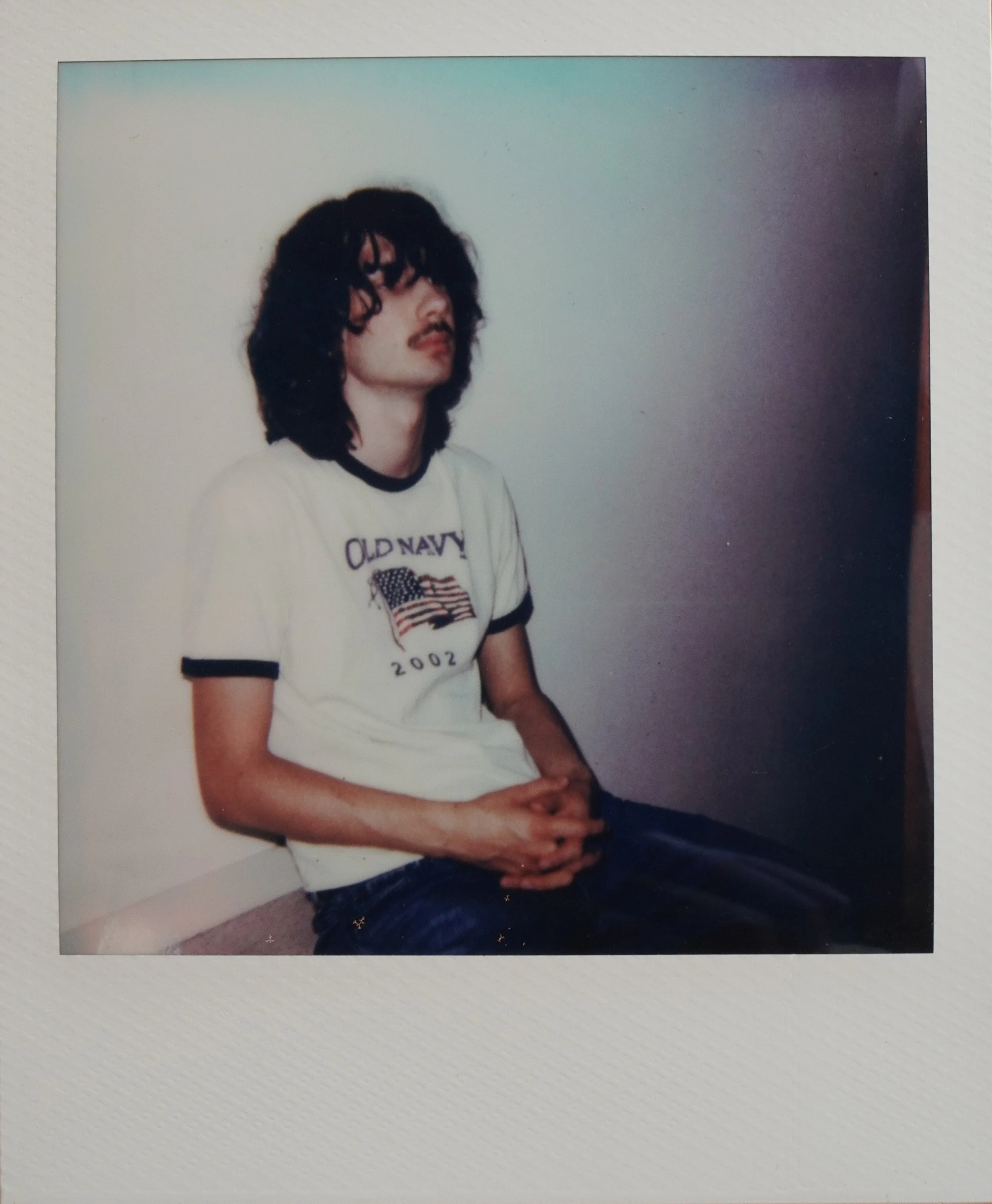 Jeune homme avec des cheveux longs et bouclés portant un t-shirt blanc Old Navy avec un drapeau américain, assis contre un mur blanc, regardant vers la gauche, mains jointes sur ses genoux.