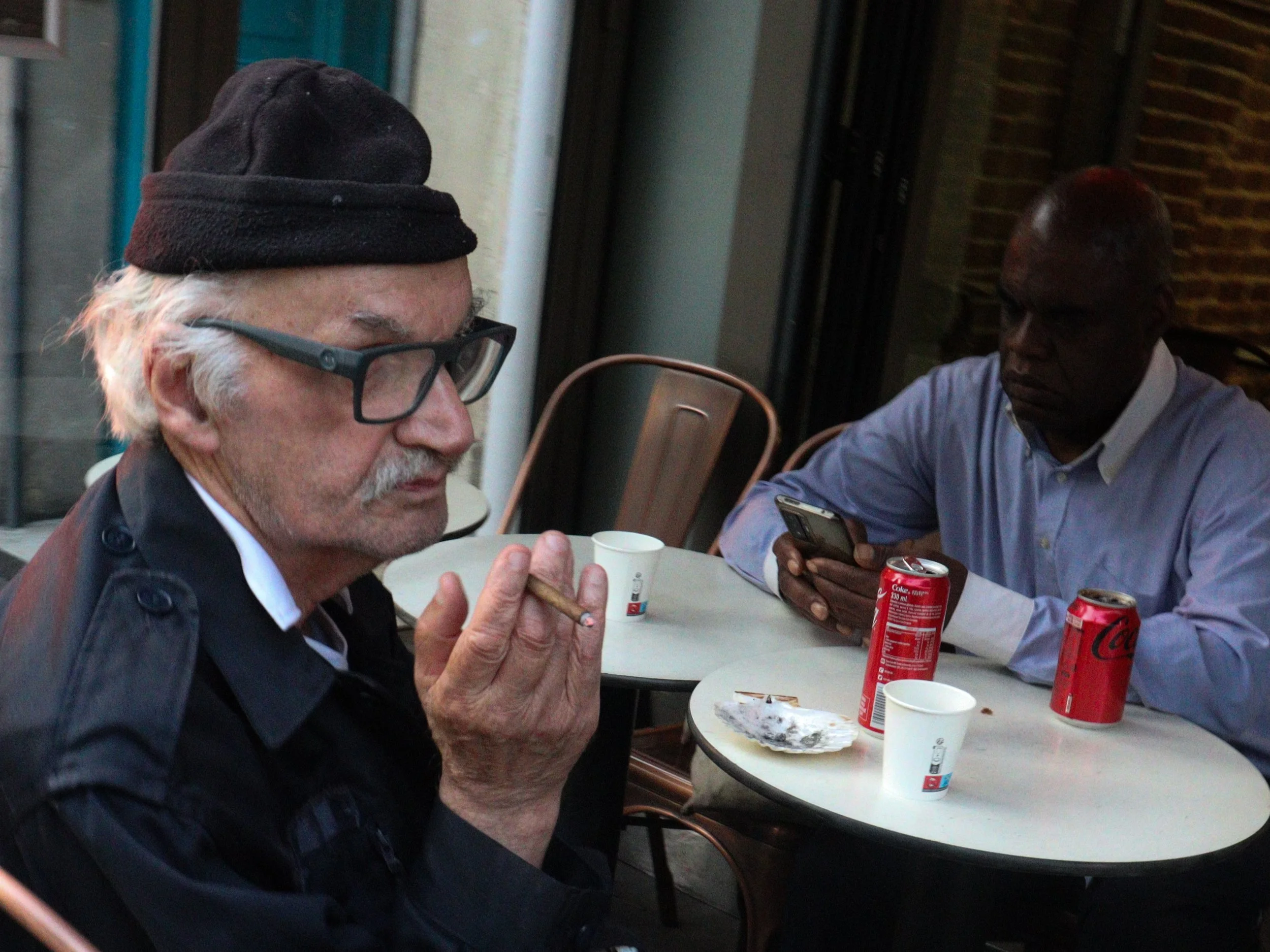 Deux hommes assis à une table de café, l'un fumant une cigarette, l'autre regardant son téléphone, avec des canettes de Coca-Cola sur la table.