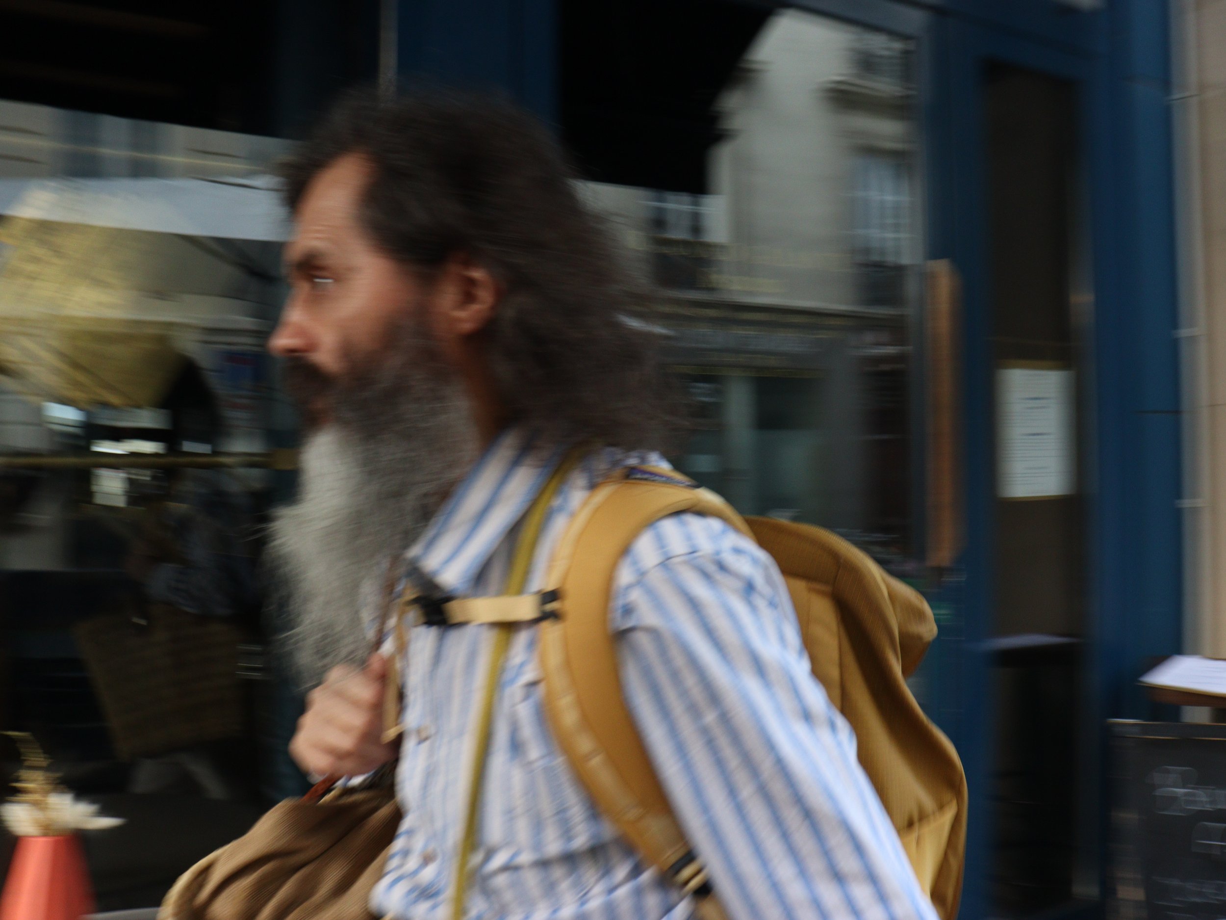 Un homme avec une longue barbe et cheveux longs portant un sac à dos beige, marchant dans une rue urbaine.