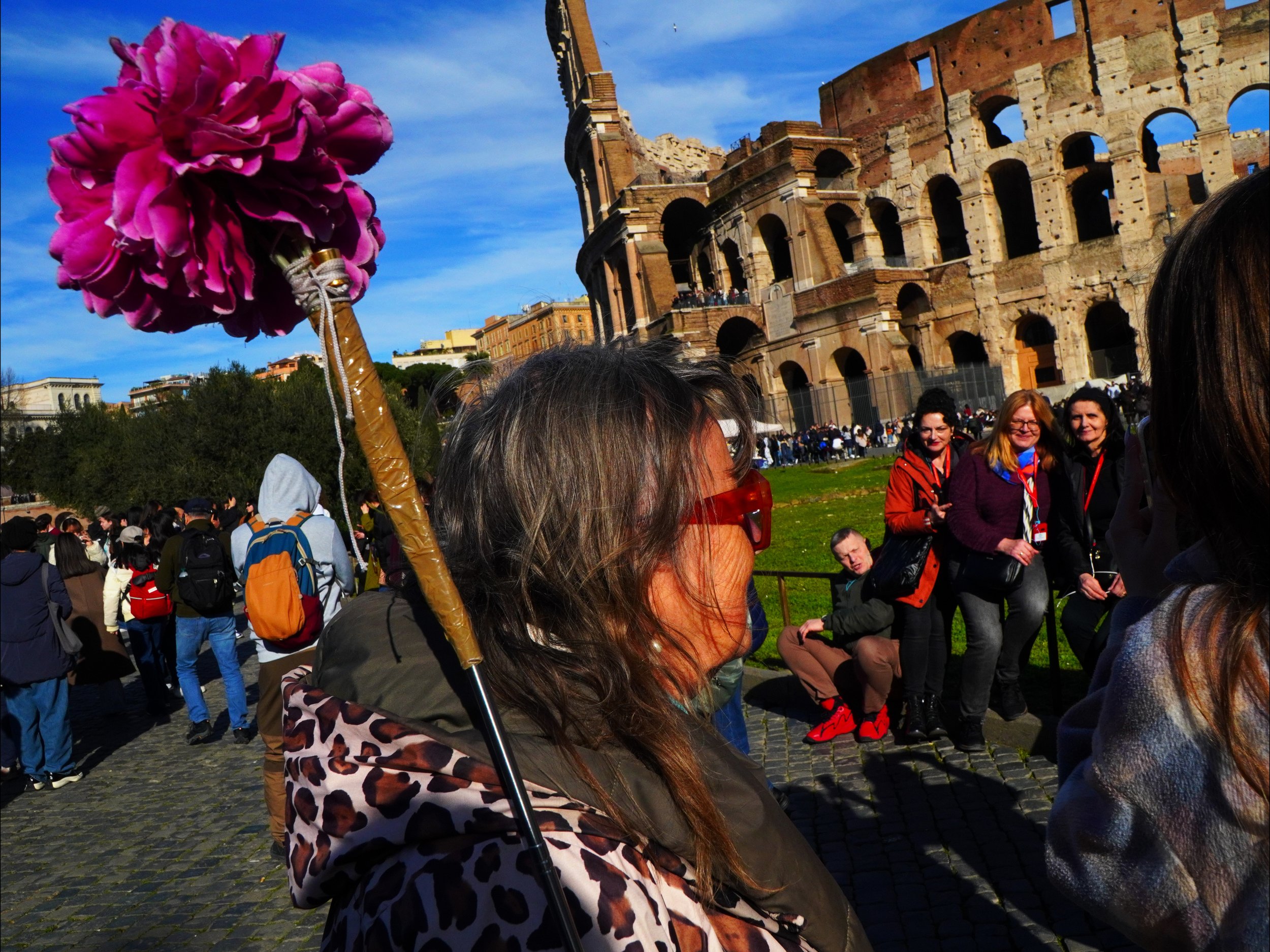 Foule de touristes devant le Colisée à Rome, avec une femme en premier plan portant un parapluie rose et des lunettes de soleil, le ciel bleu en arrière-plan.