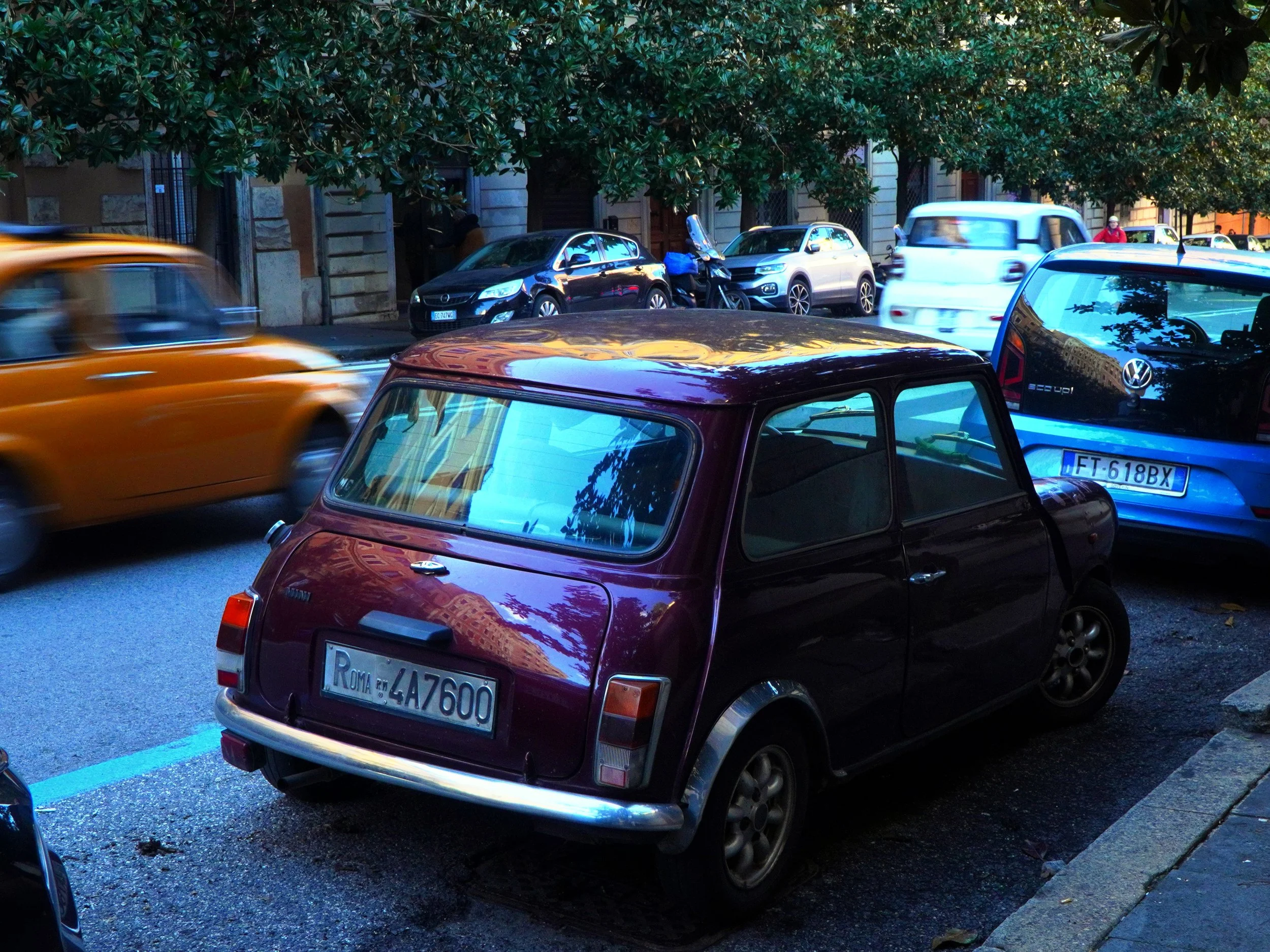 Une petite voiture ancienne noire avec une plaque d'immatriculation romaine, stationnée sur le côté d'une rue animée avec plusieurs autres voitures modernes qui passent rapidement.
